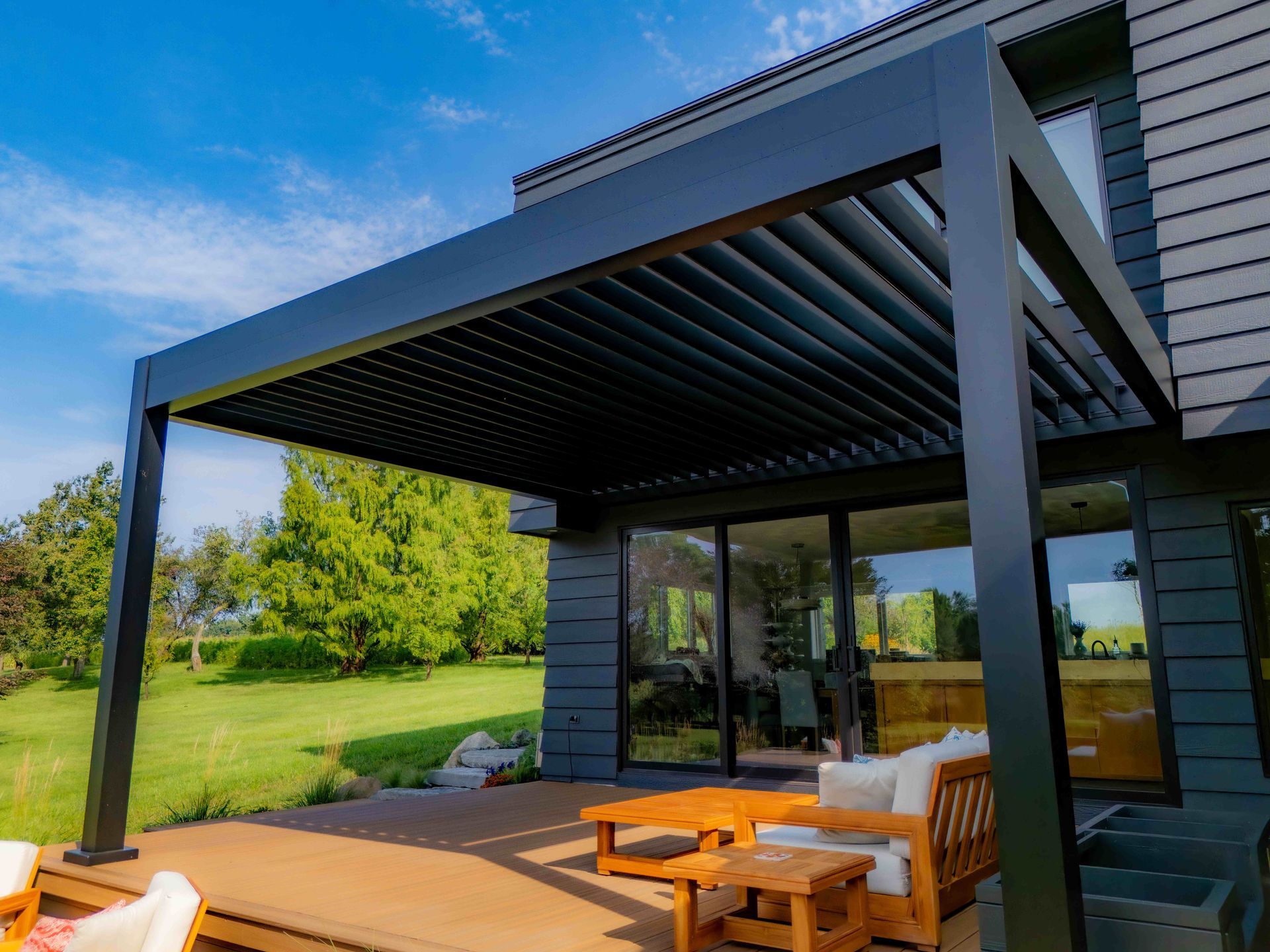 A modern black louvered pergola extends from a dark-sided house over a wooden patio with outdoor furniture and a lawn.