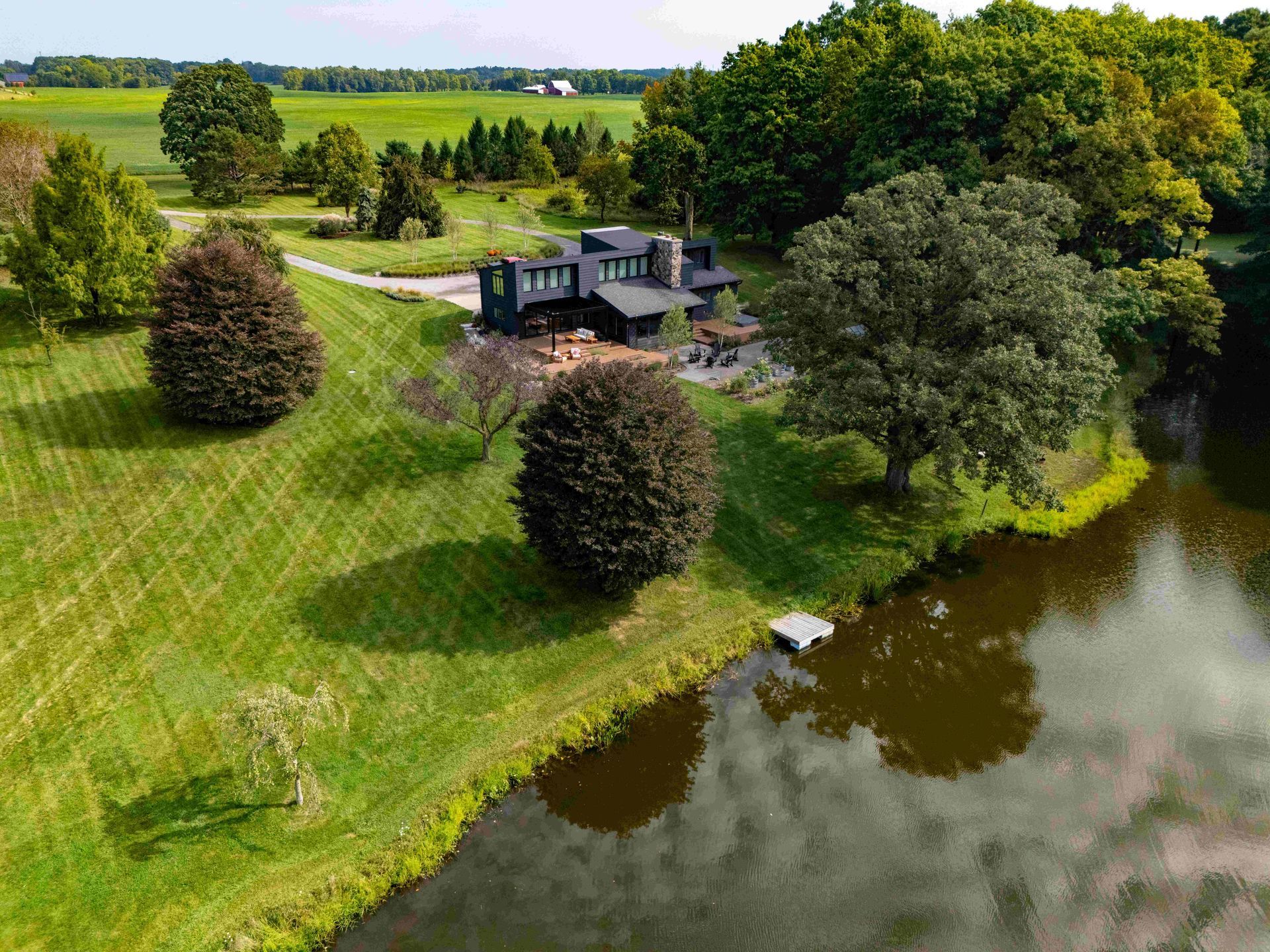 An aerial view of a dark-colored, modern house situated on a grassy hill next to a pond and dense woods.