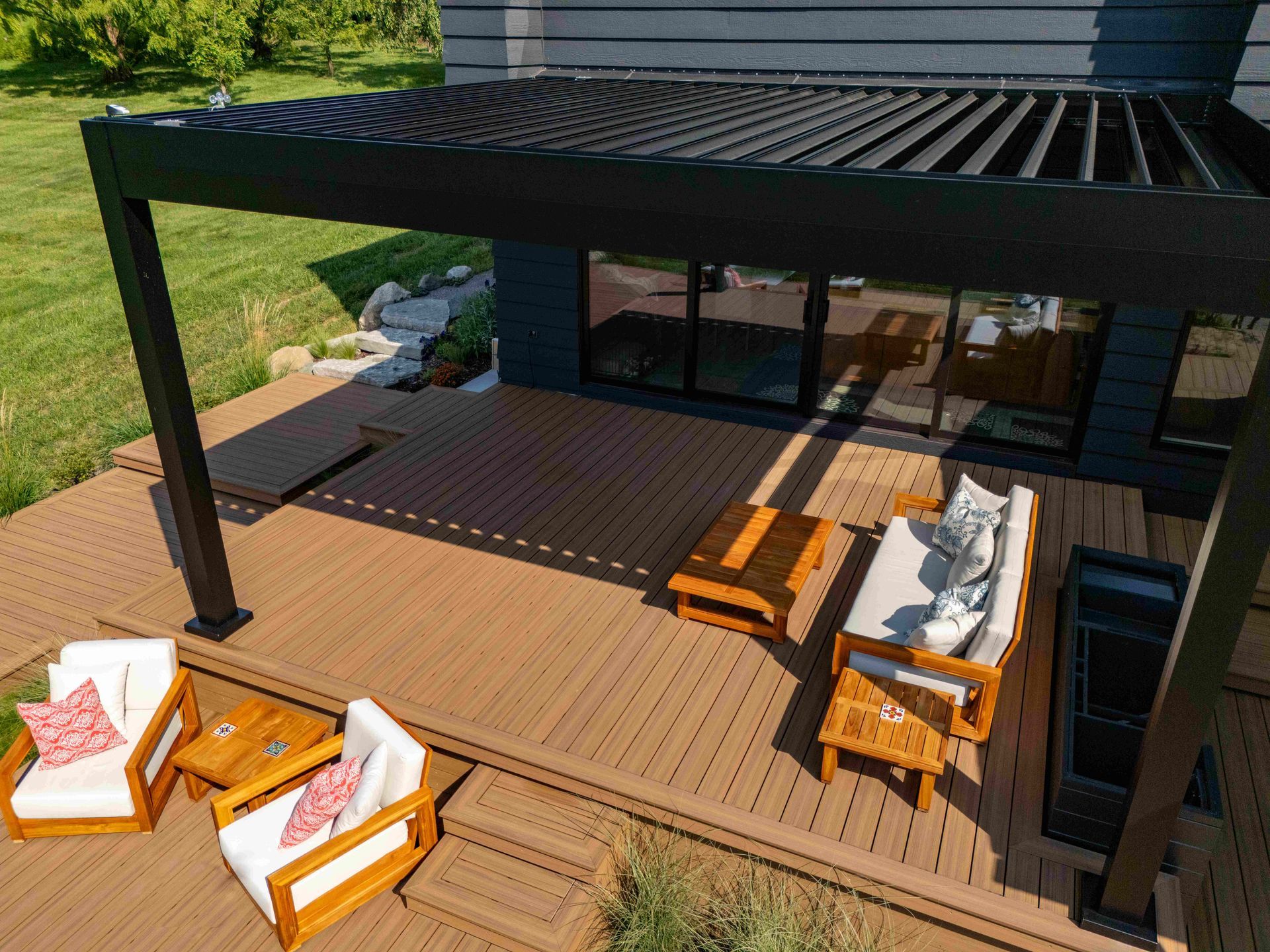 An elevated, high-angle view of a wooden patio deck featuring a black louvered pergola, outdoor seating, and a table.