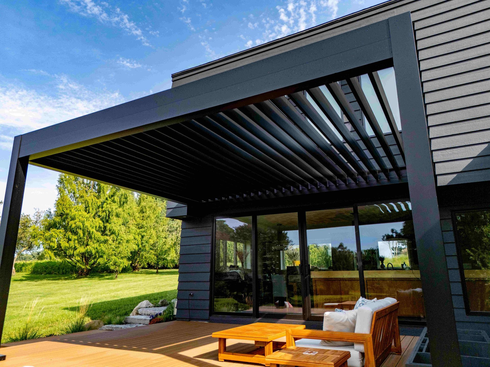 A modern home exterior with a black louvered pergola covering a wooden deck, sliding glass doors, and a grassy backyard.
