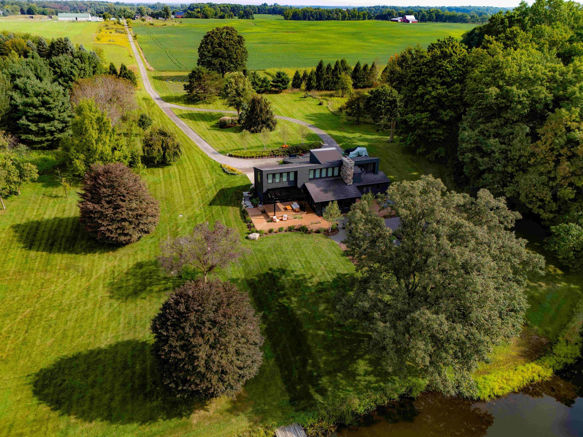 An aerial view of a dark-sided house with a stone patio, nestled in a grassy landscape with trees and a nearby pond.