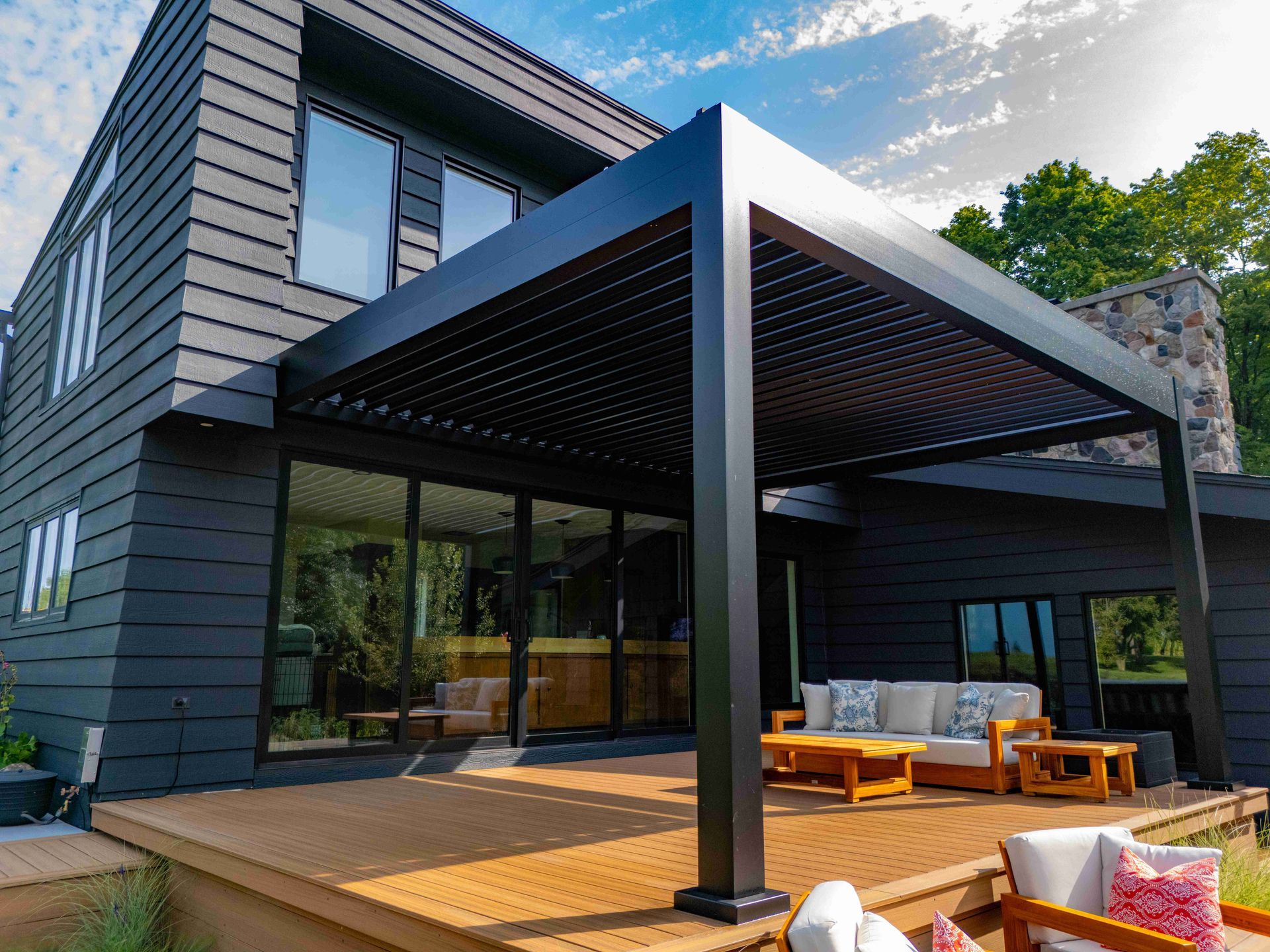 Modern home exterior featuring a black louvered pergola over a wooden deck with outdoor seating.