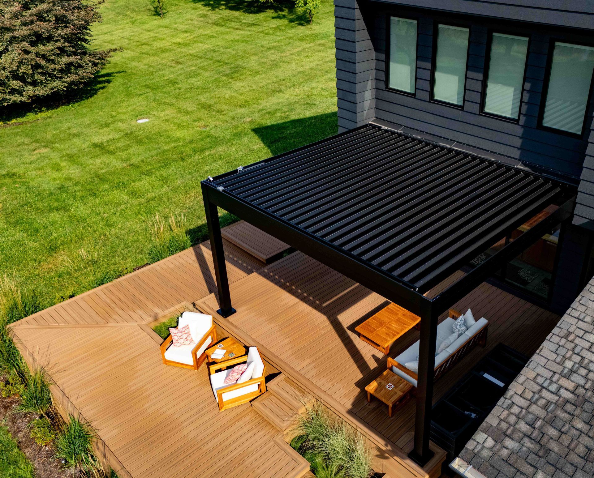 A black louvered pergola stands on a brown wooden deck outside a house with gray siding, featuring patio seating.