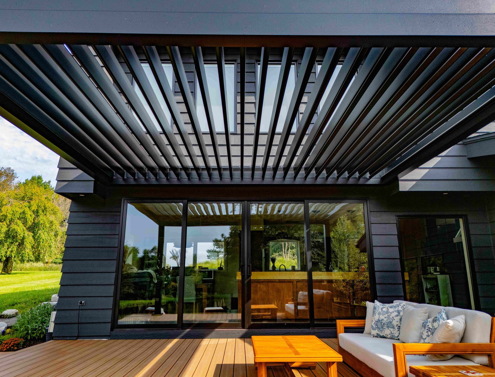 Modern patio with a dark louvered pergola roof, outdoor seating, and a wooden deck overlooking a grassy lawn.