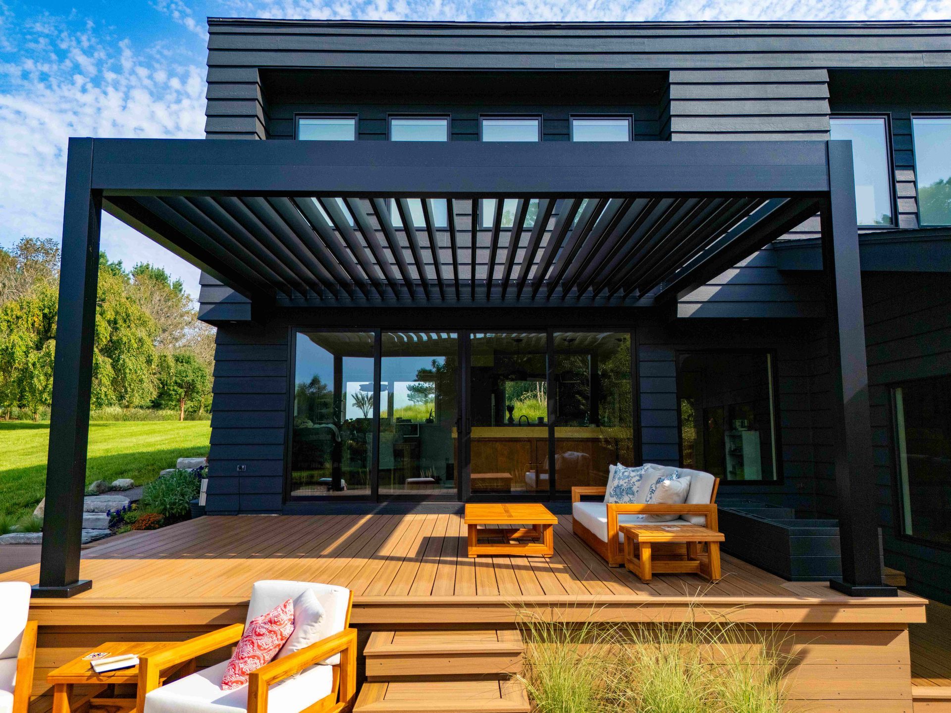 A modern, black-framed pergola covers a wooden deck with outdoor seating in front of a dark, contemporary house.