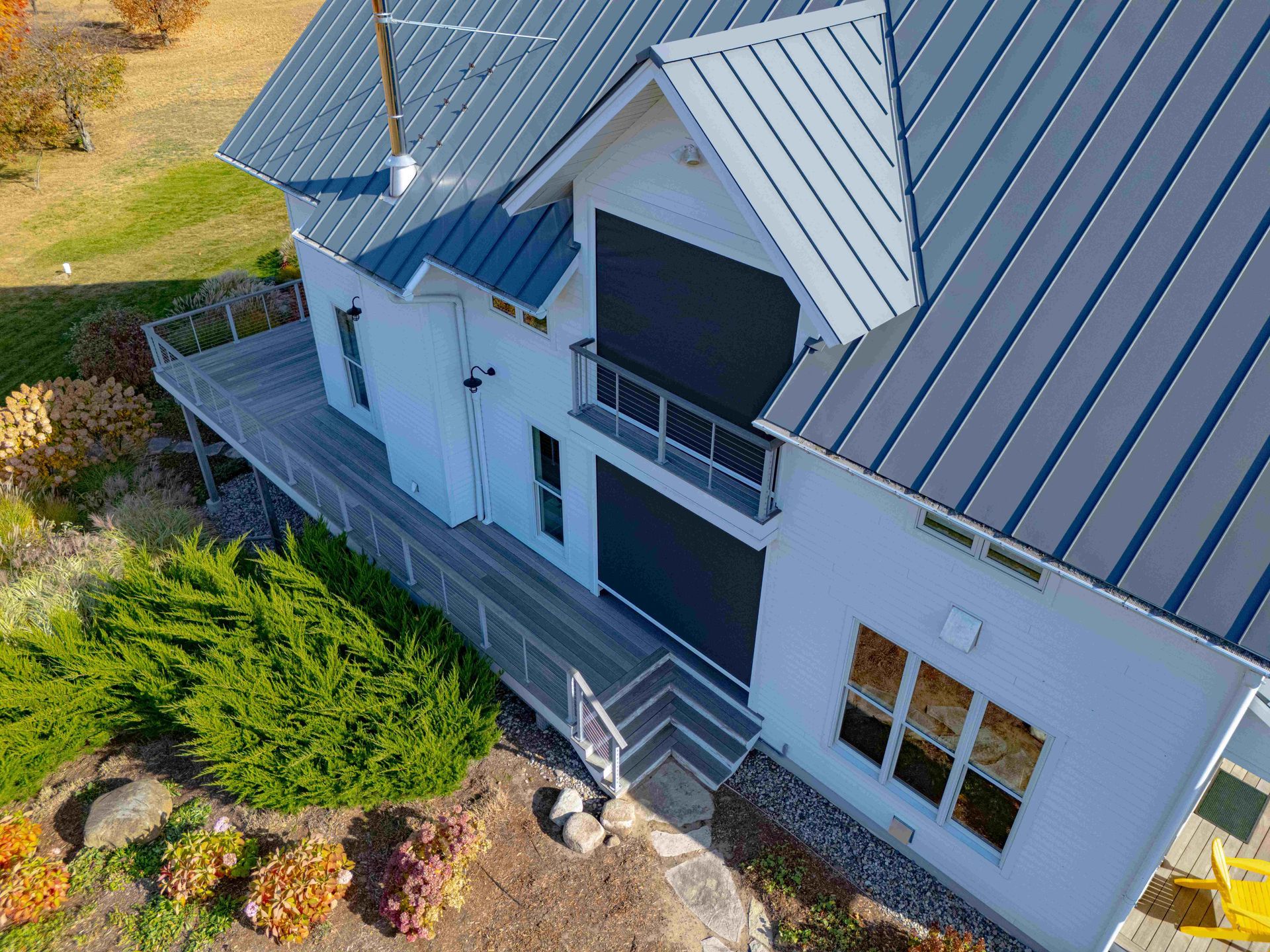 High-angle view of a white house with a grey metal roof and deck, surrounded by landscaping.
