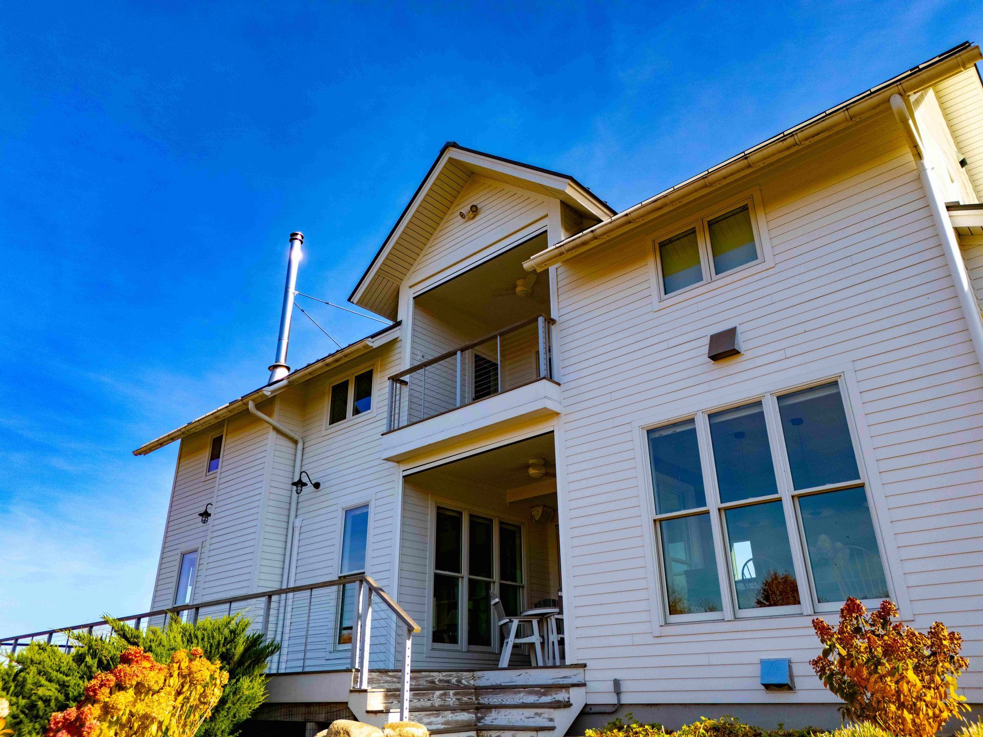 A two-story white house with a balcony, large windows, and a metal chimney against a clear blue sky.