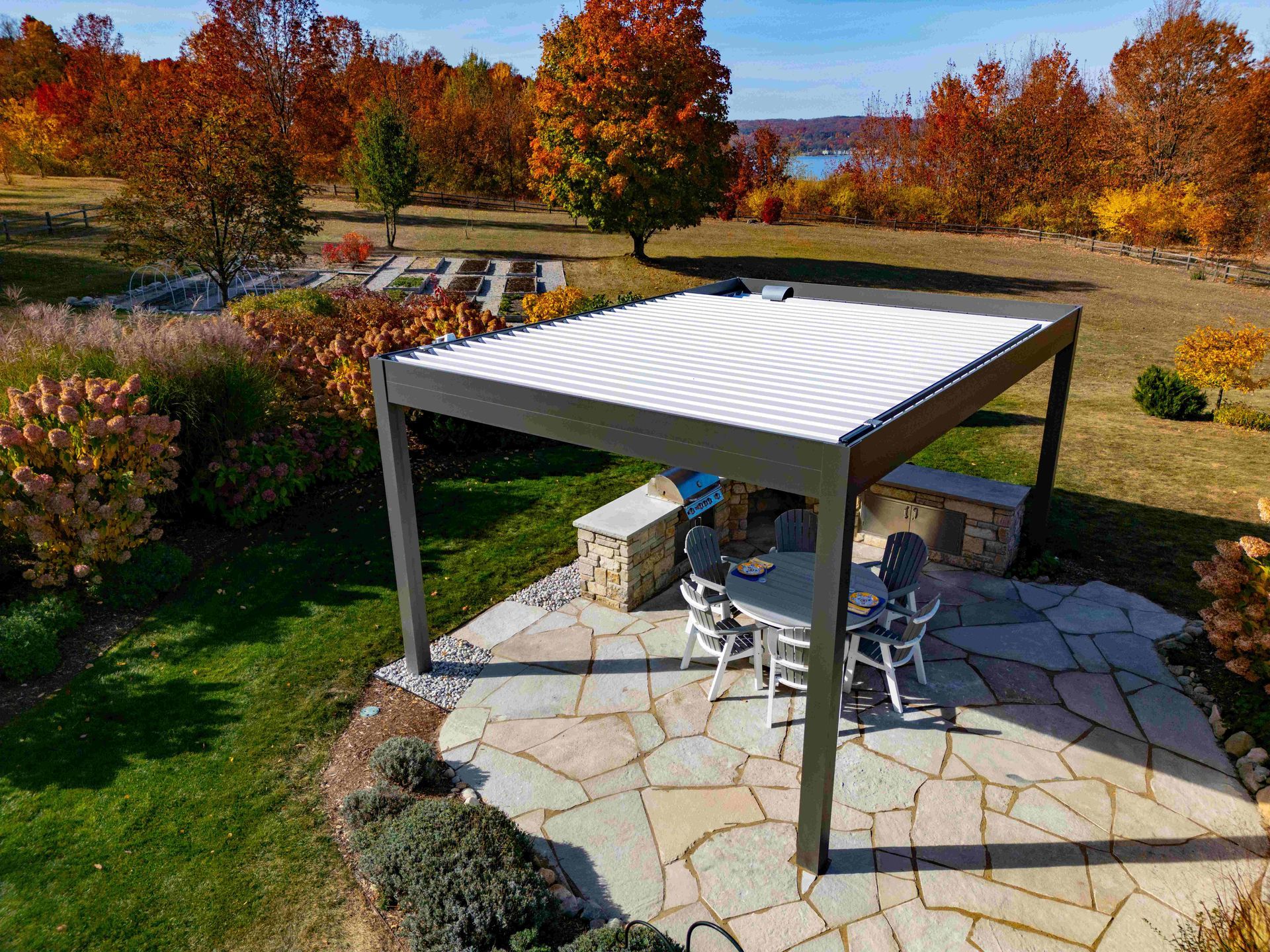 A modern, dark gray pergola with a white retractable roof covering a stone patio dining area on a sunny autumn day.