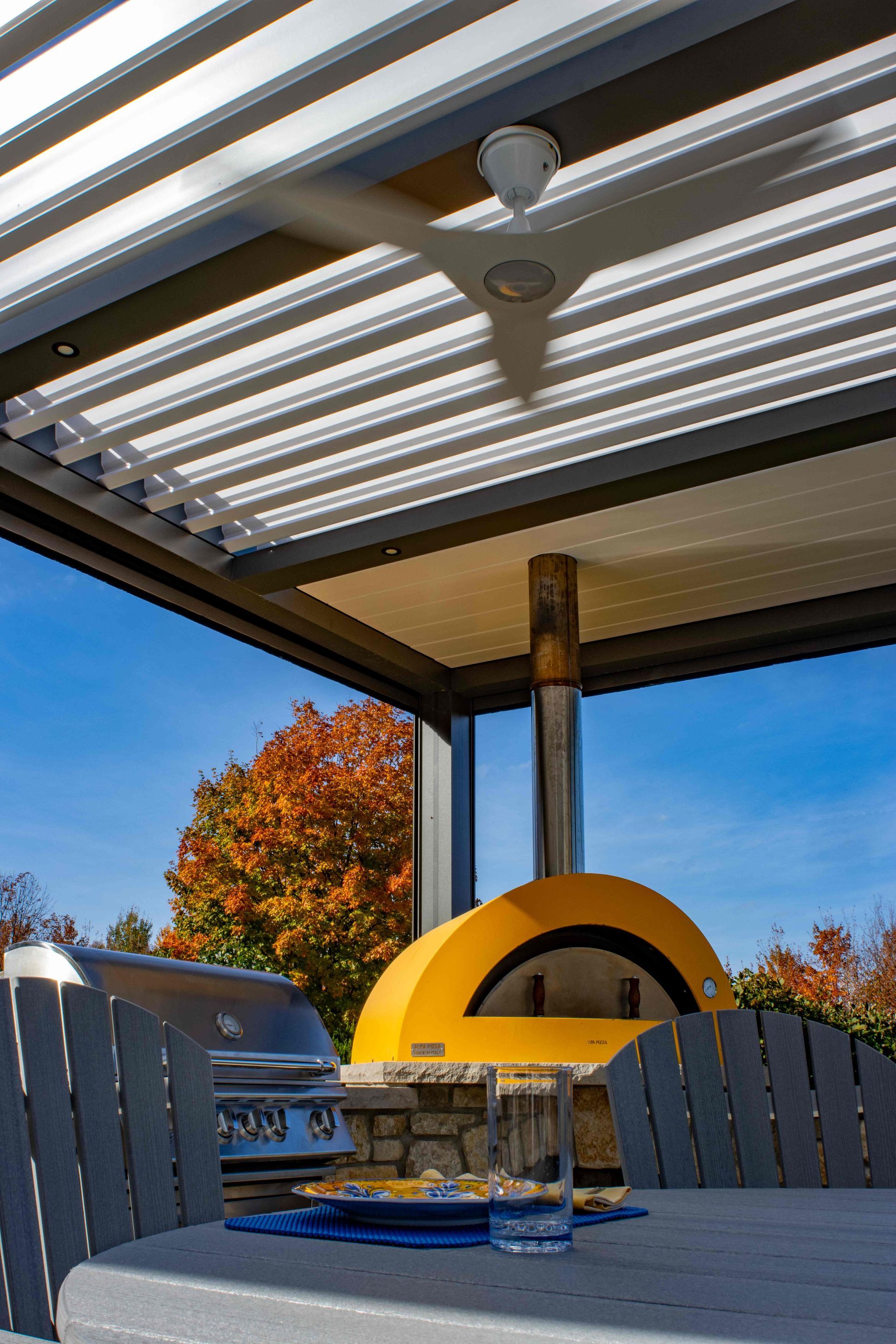 Outdoor patio with a yellow pizza oven, a grill, seating, and a slatted roof under a clear blue sky.