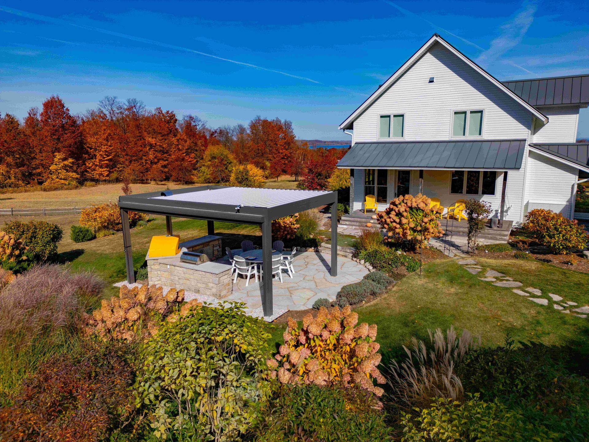 A white farmhouse with a stone patio, outdoor kitchen, and pergola surrounded by autumn trees under a bright blue sky.