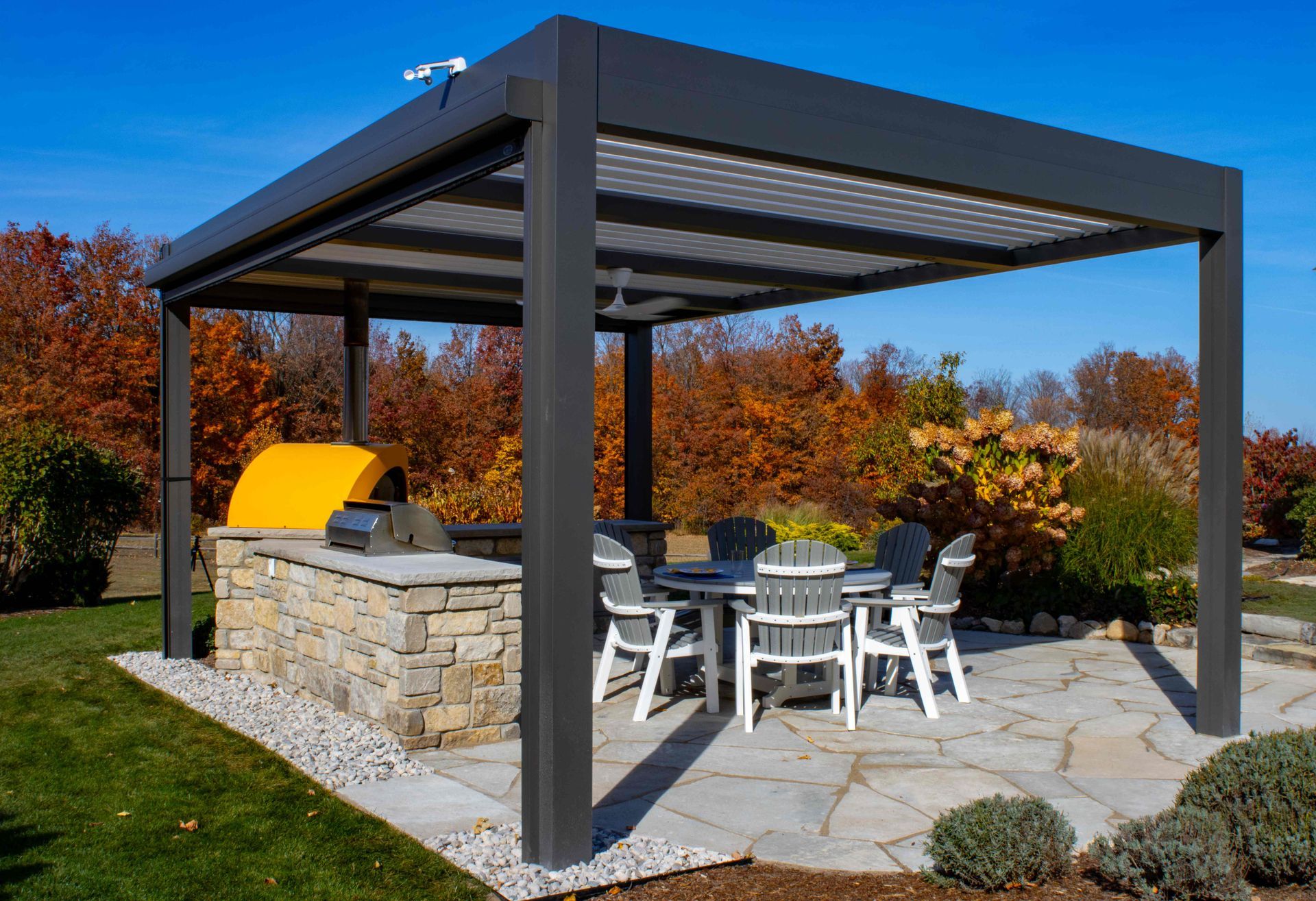 A covered outdoor patio featuring a stone kitchen with a yellow pizza oven and a dining set on a flagstone floor.