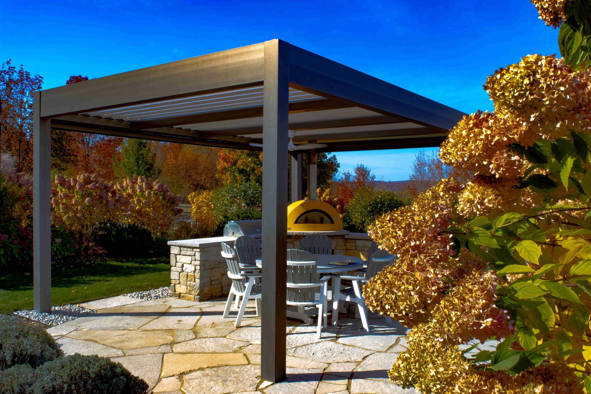 A modern metal pergola over a stone patio with outdoor dining furniture, a pizza oven, and golden fall foliage nearby.