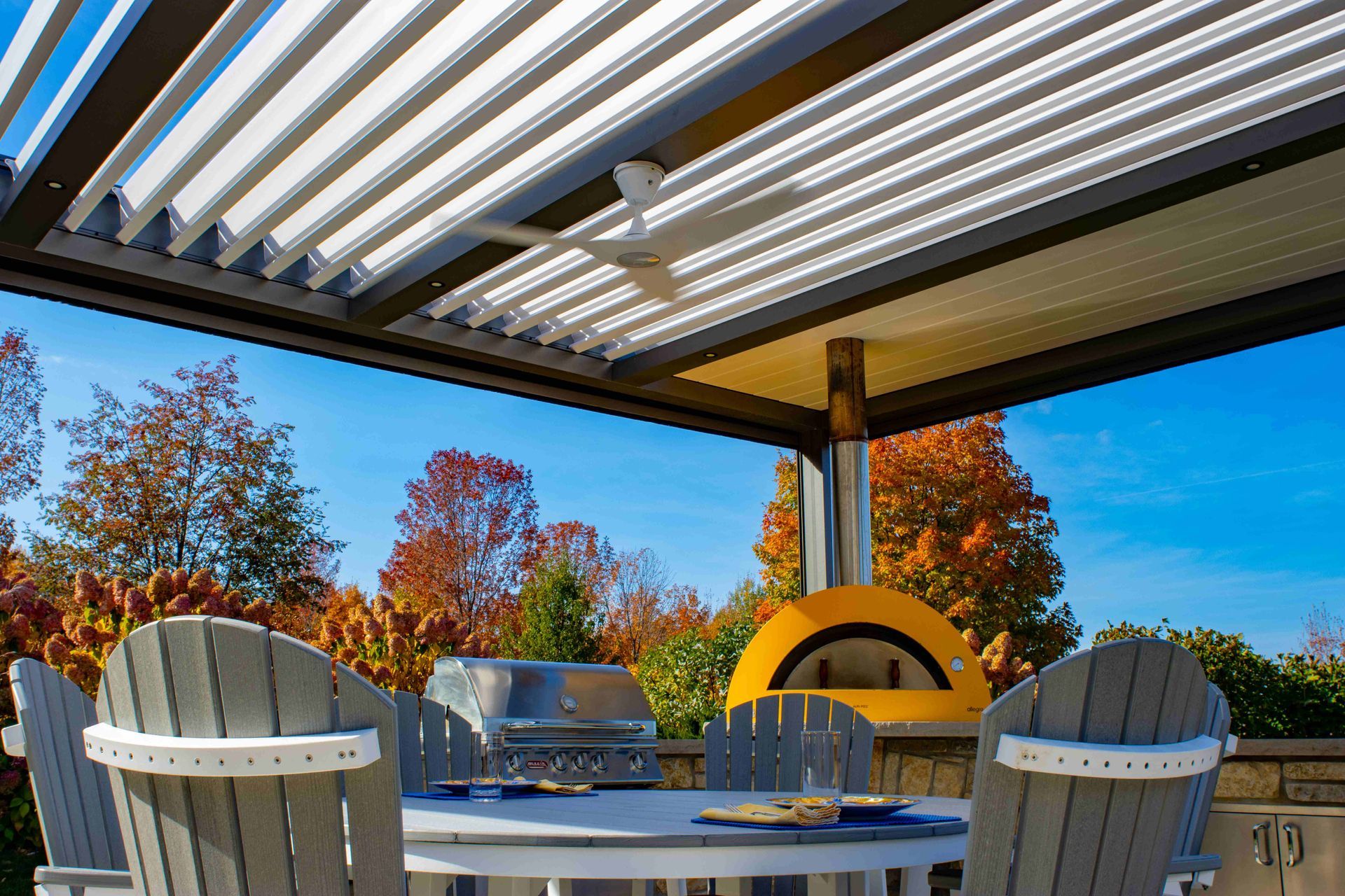 Outdoor patio with a pergola, dining table, chairs, grill, and a yellow pizza oven set against an autumn forest.