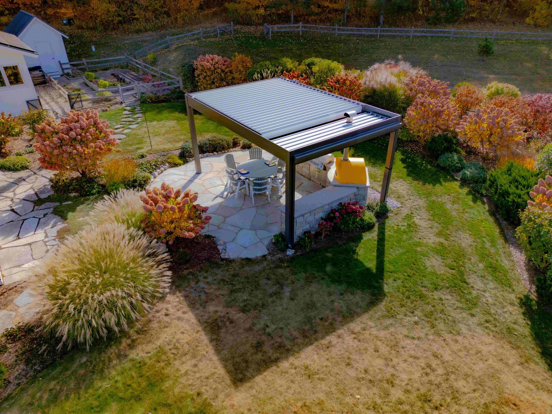 A pergola with a louvered roof sits on a stone patio in a backyard garden during autumn, surrounded by colorful foliage.