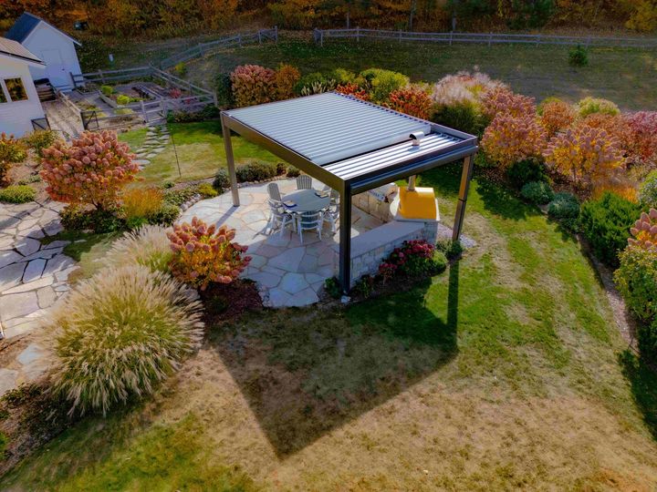 A pergola with a louvered roof sits on a stone patio in a backyard garden during autumn, surrounded by colorful foliage.