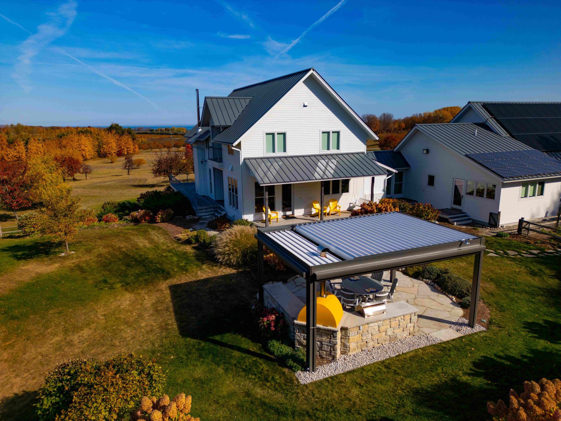 A white two-story house with a grey roof and a detached patio pergola, situated in a field during autumn.