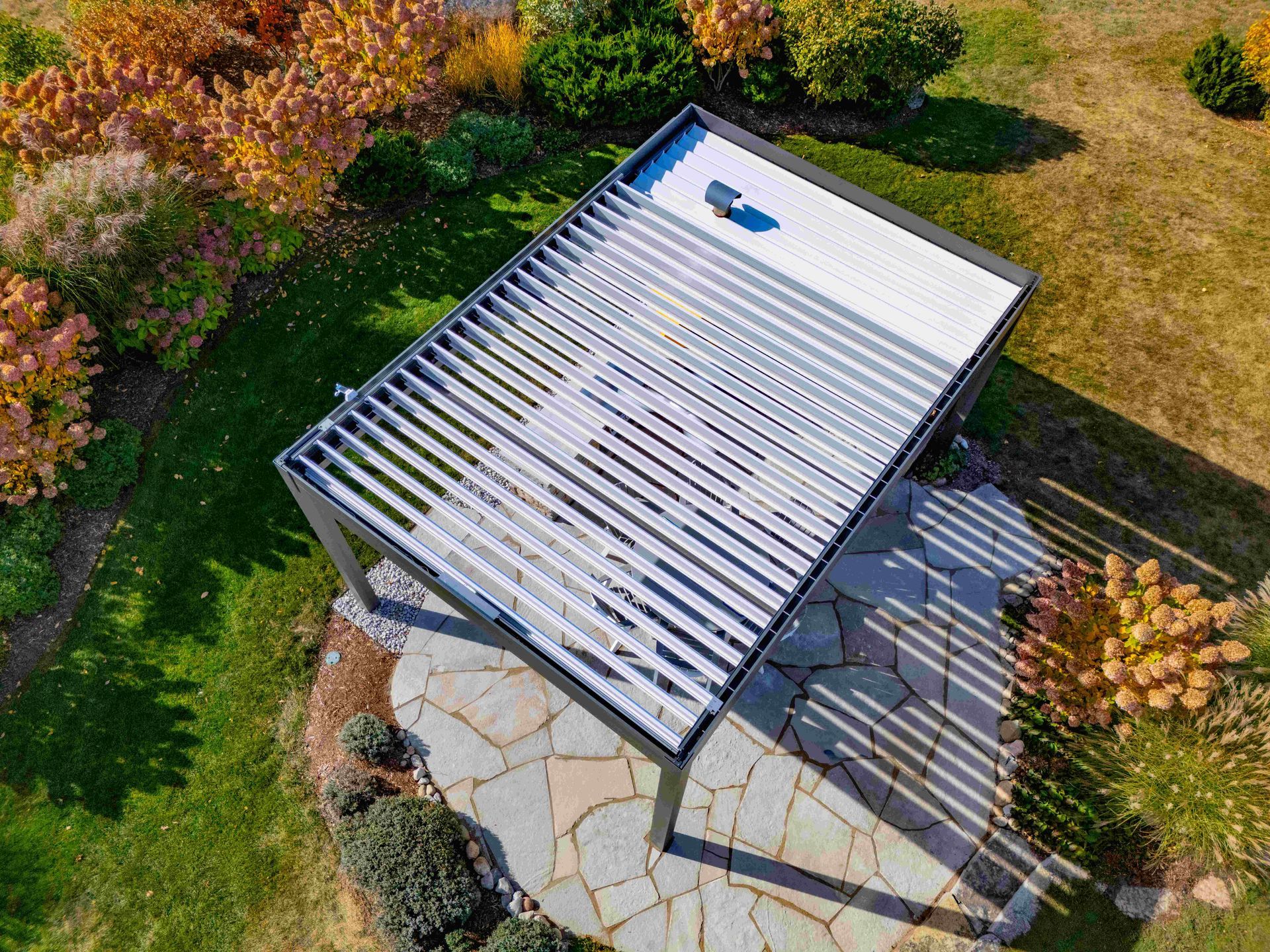 High-angle view of a patio pergola with adjustable silver louvers, installed over a flagstone patio in a sunny garden.