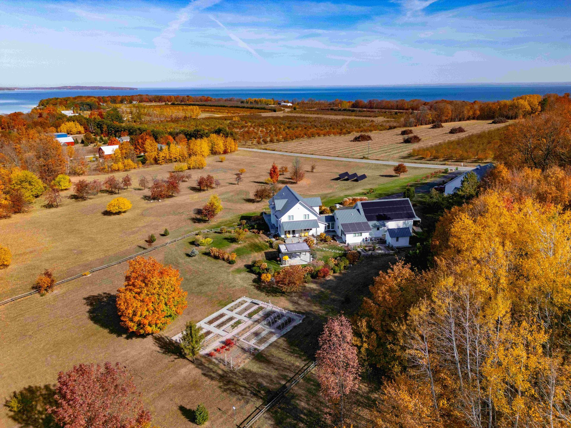 Aerial view of a white farmhouse and garden surrounded by autumn trees and fields, with a body of water in the distance.