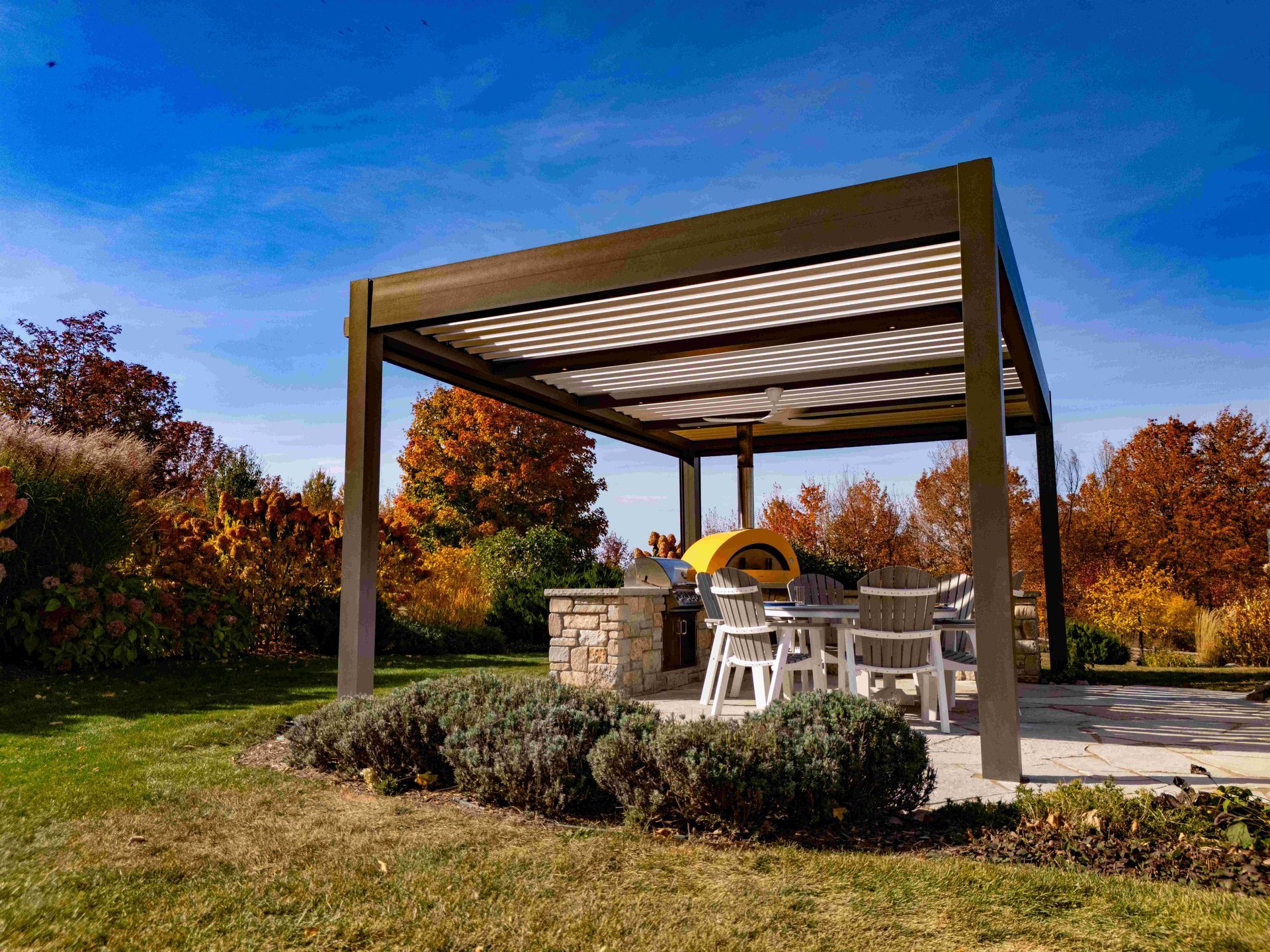 A pergola with a louvered roof covers a stone outdoor kitchen and dining area in a backyard during autumn.