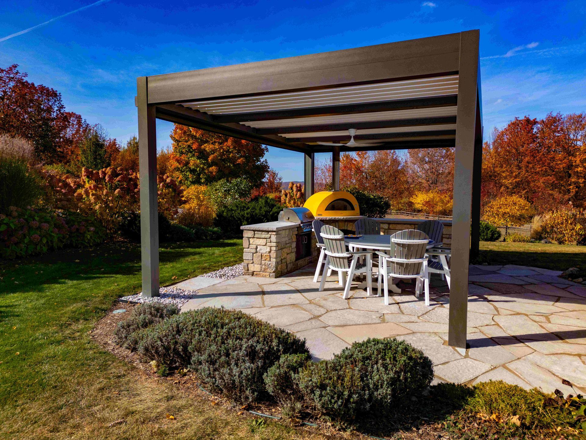 A metal pergola covers a stone patio with an outdoor kitchen, a yellow pizza oven, and a dining set amid autumn foliage.