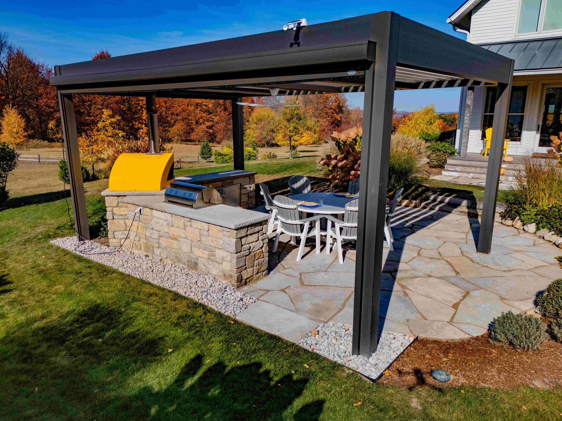 A stone outdoor kitchen with a yellow grill under a dark pergola, next to a dining set on a flagstone patio.