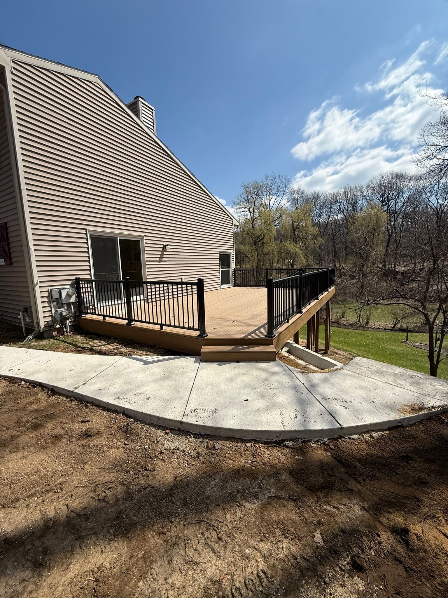 A beige house features a new wooden deck with black railings and a curved concrete walkway leading to the backyard.