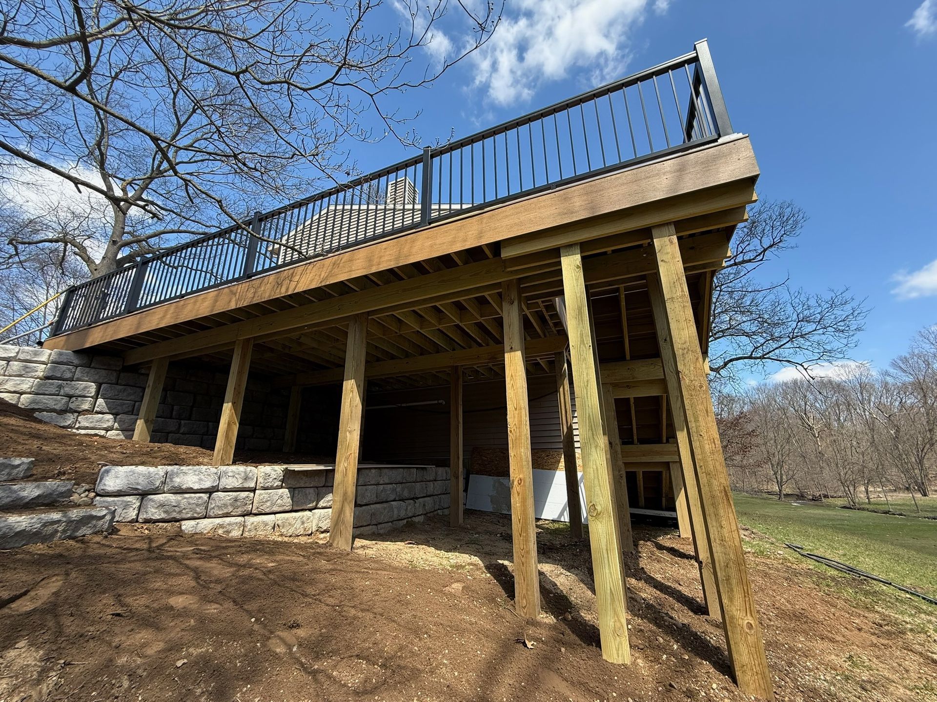 A wooden deck with a black railing, supported by vertical posts, overlooking a stone retaining wall and grassy yard.