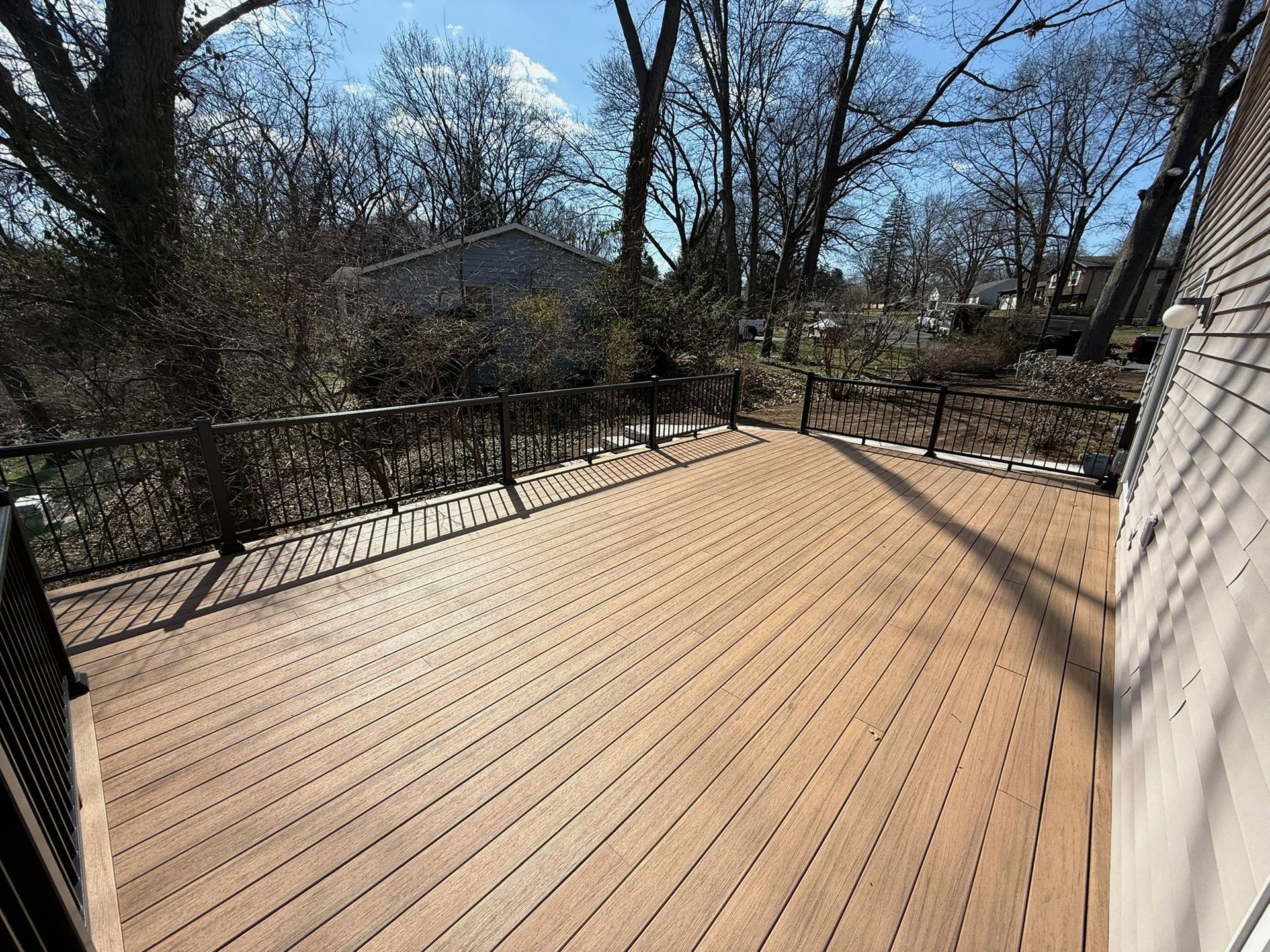 A wooden deck with a black railing, overlooking bare trees on a sunny day.