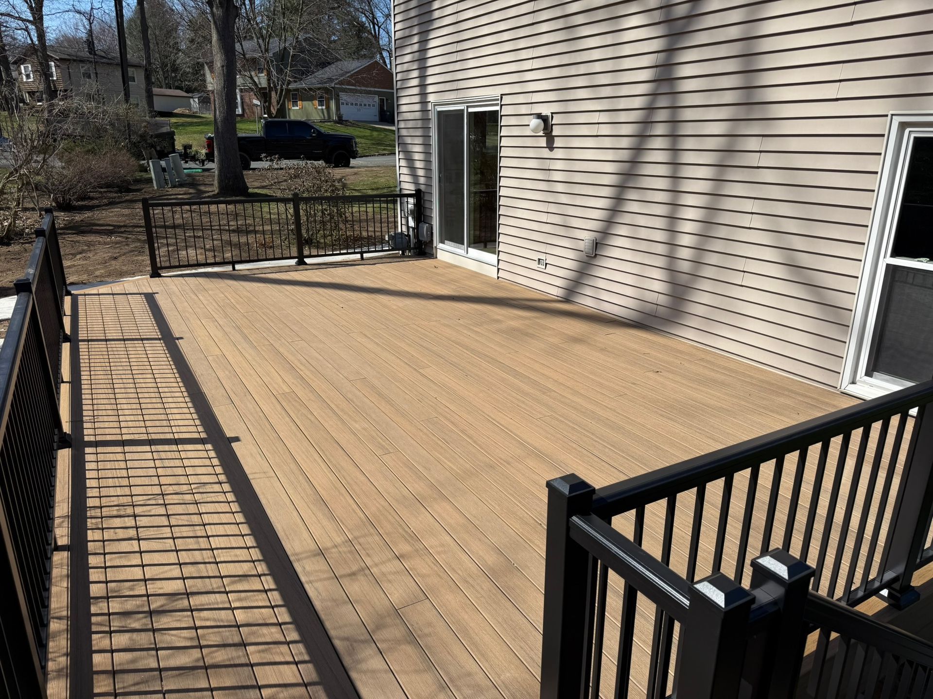 A tan wooden deck with black railings attached to a house with beige siding on a sunny day.
