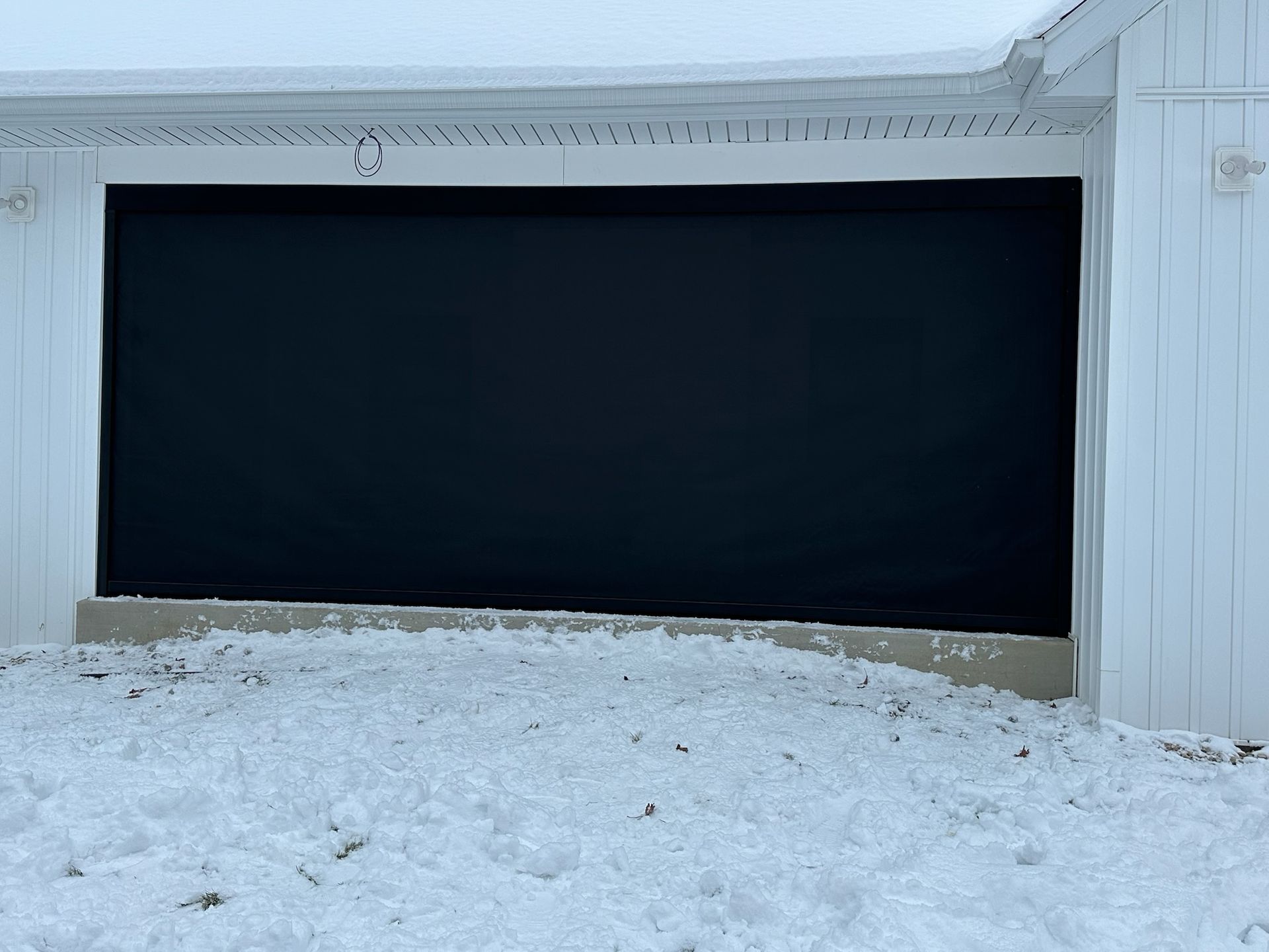 A black garage screen enclosure is installed over a white garage door opening, with snow on the ground in front.