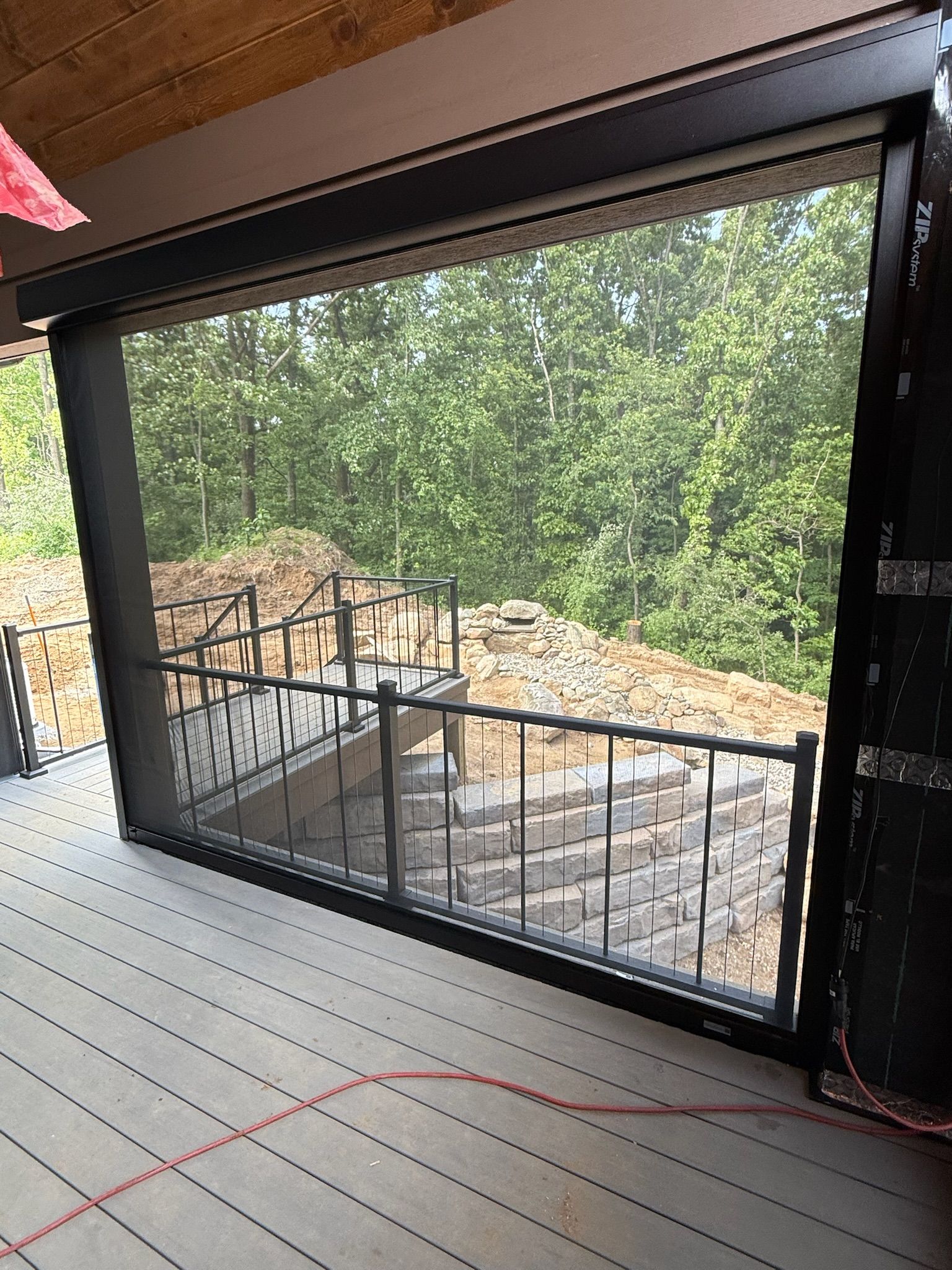 A black, motorized outdoor screen is lowered over a patio deck, partially obscuring a view of trees and stone steps.