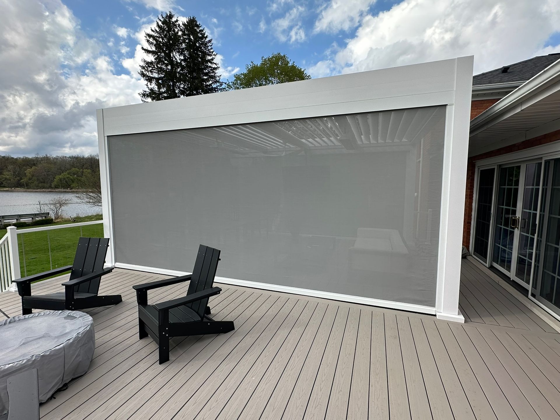A white outdoor pergola with a grey privacy screen, two black chairs, and a covered fire pit on a wooden deck by a lake.