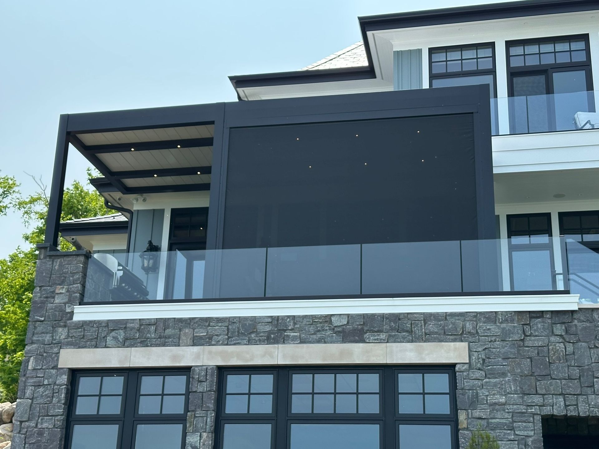 A house balcony featuring a black louvered pergola roof, integrated screen walls, and glass railings above a stone facade.