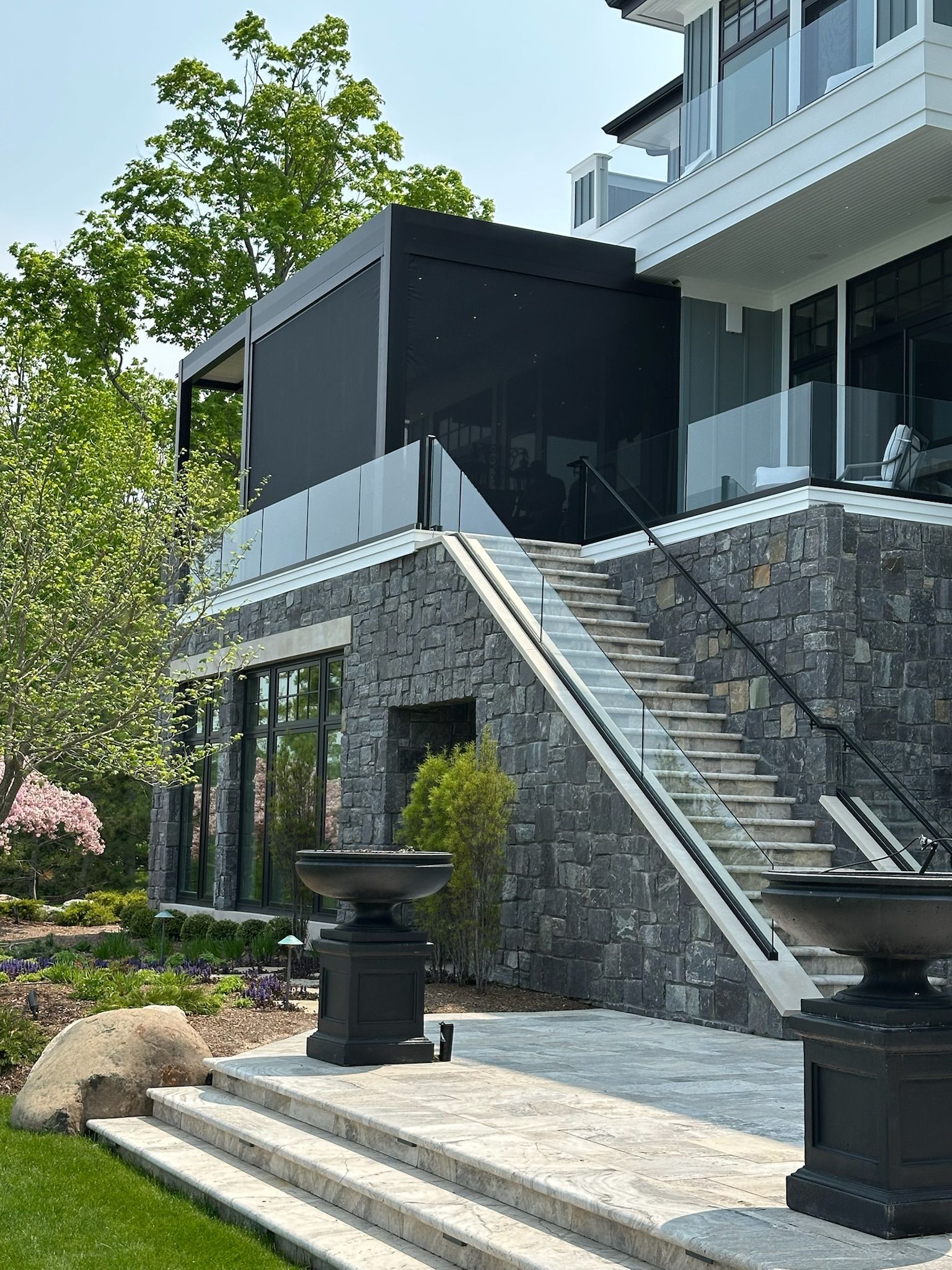 Exterior view of a multi-level stone house with a modern deck, glass railings, and a black privacy screen.