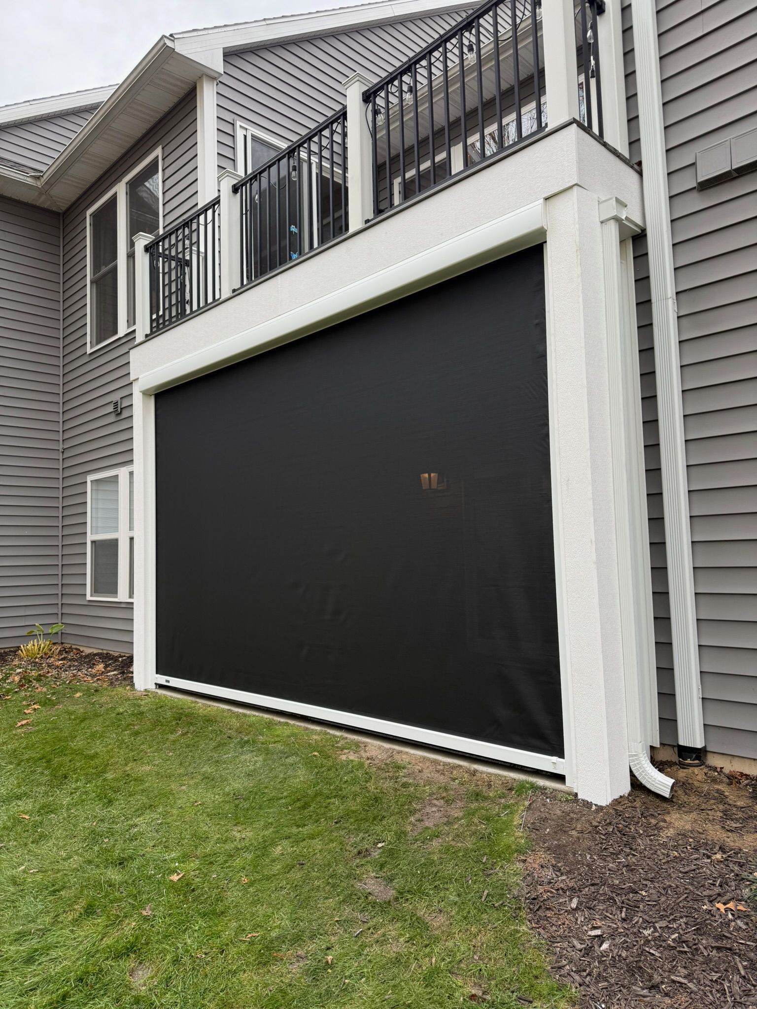A black privacy shade installed under a deck with white wooden support beams against a grey siding house.