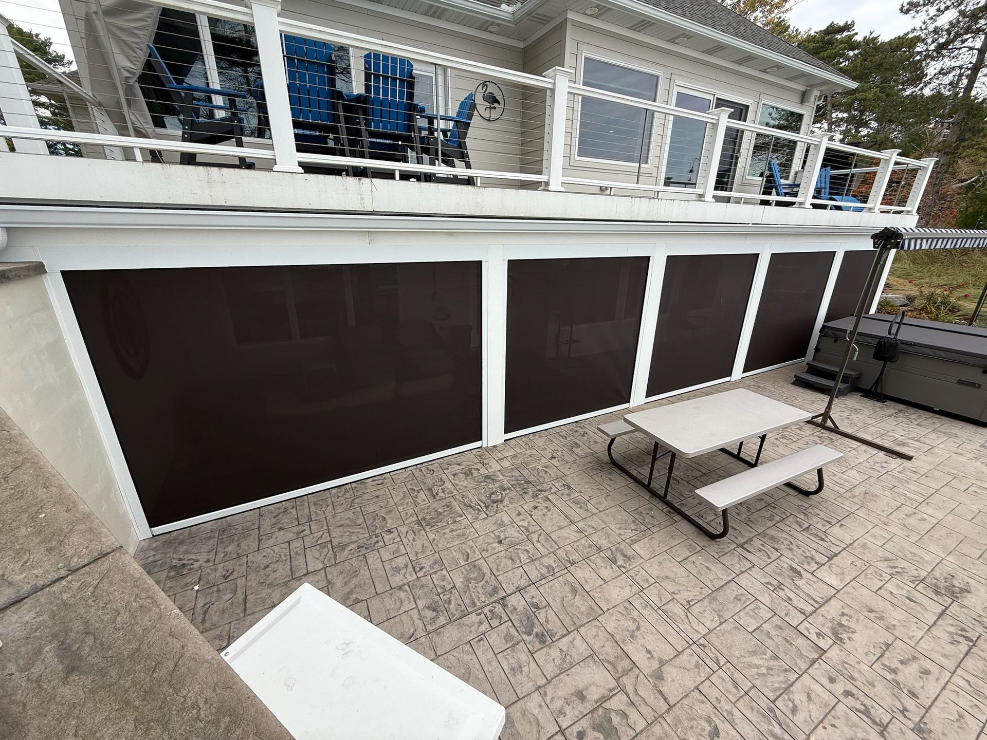 A ground-level patio with a stone-textured floor, a picnic table, and a deck above enclosed by dark privacy screens.