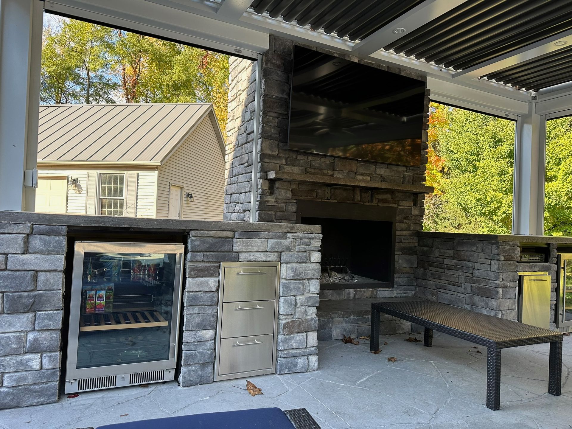 A stone outdoor kitchen featuring a built-in refrigerator, cabinet, fireplace, and mounted television under a pergola.