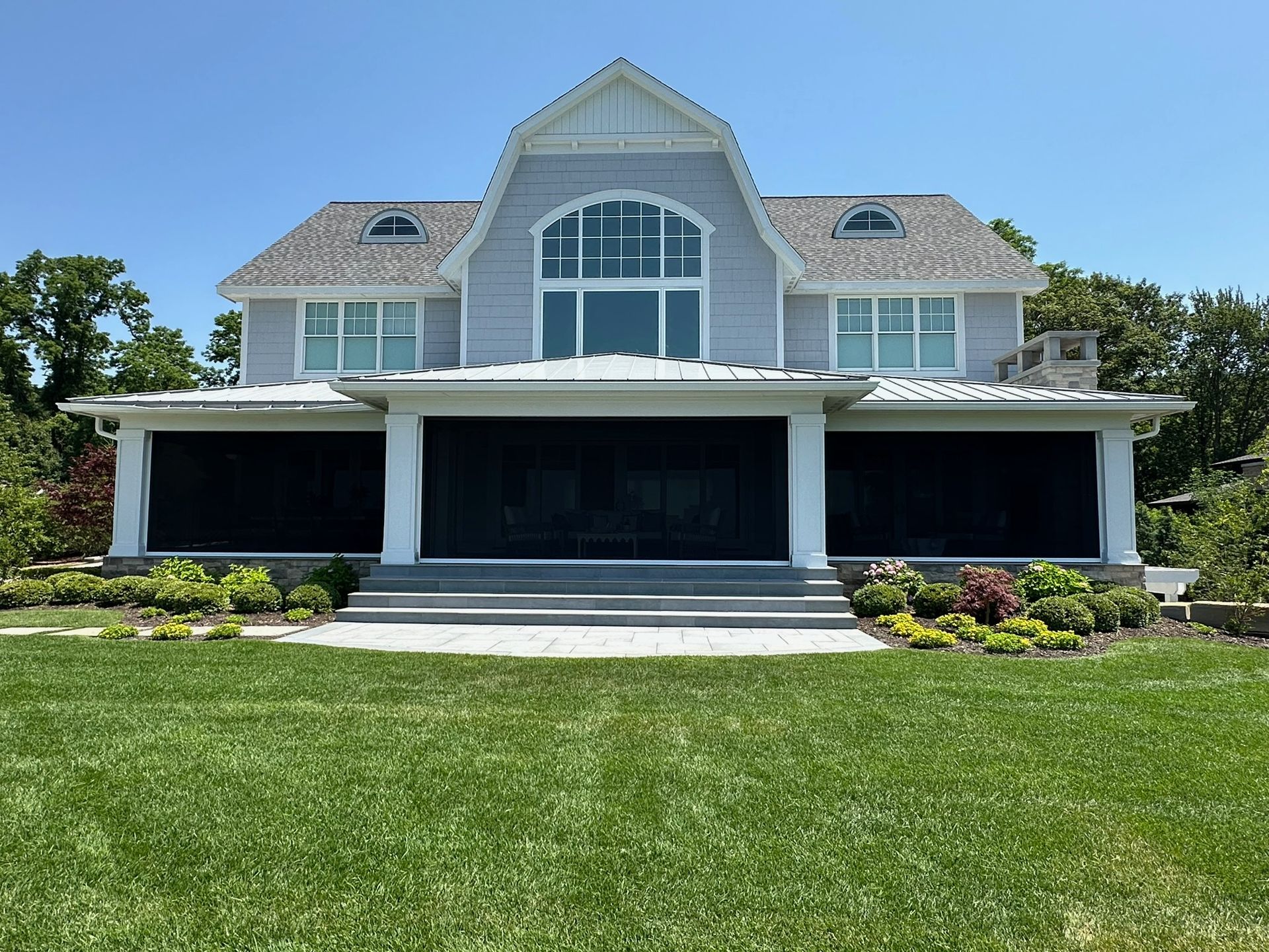 A gray, gambrel-roofed house with a large covered porch and black privacy screens, set behind a lush green lawn.