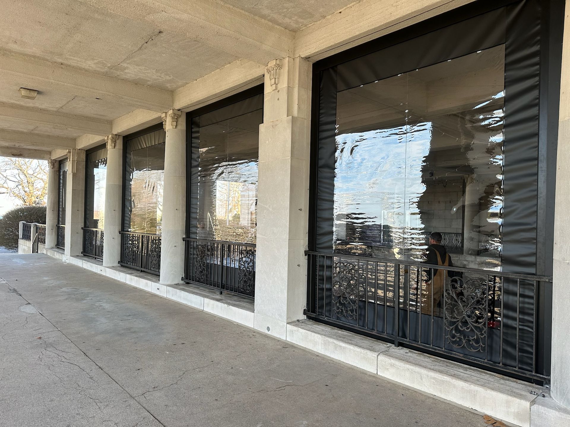 Outdoor patio with clear, heavy-duty vinyl weather curtains attached to stone pillars and an iron railing.