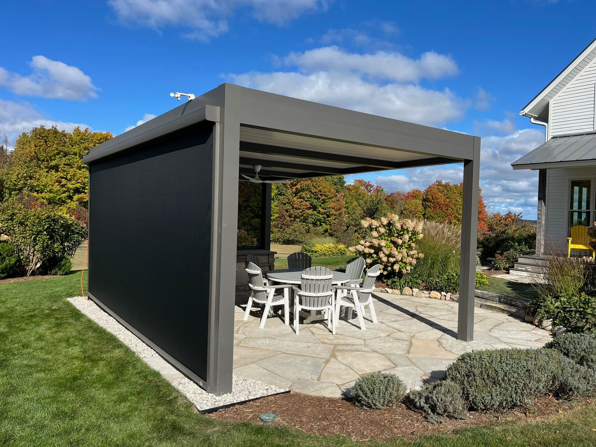A modern dark gray pergola with a retractable privacy screen set on a stone patio in a backyard on a sunny day.