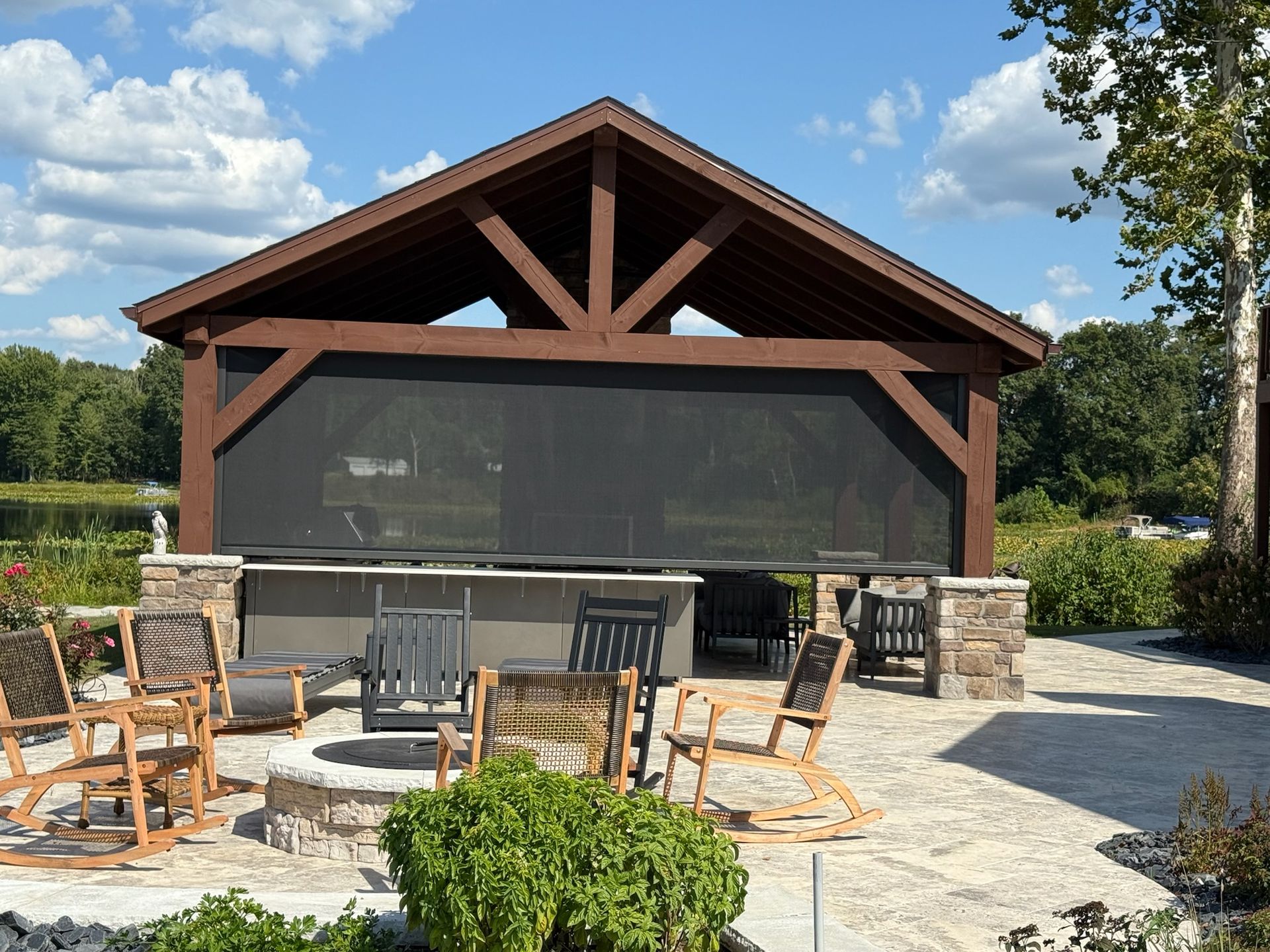 A timber-framed outdoor pavilion with stone pillars, a fire pit, and rocking chairs on a stone patio under a blue sky.