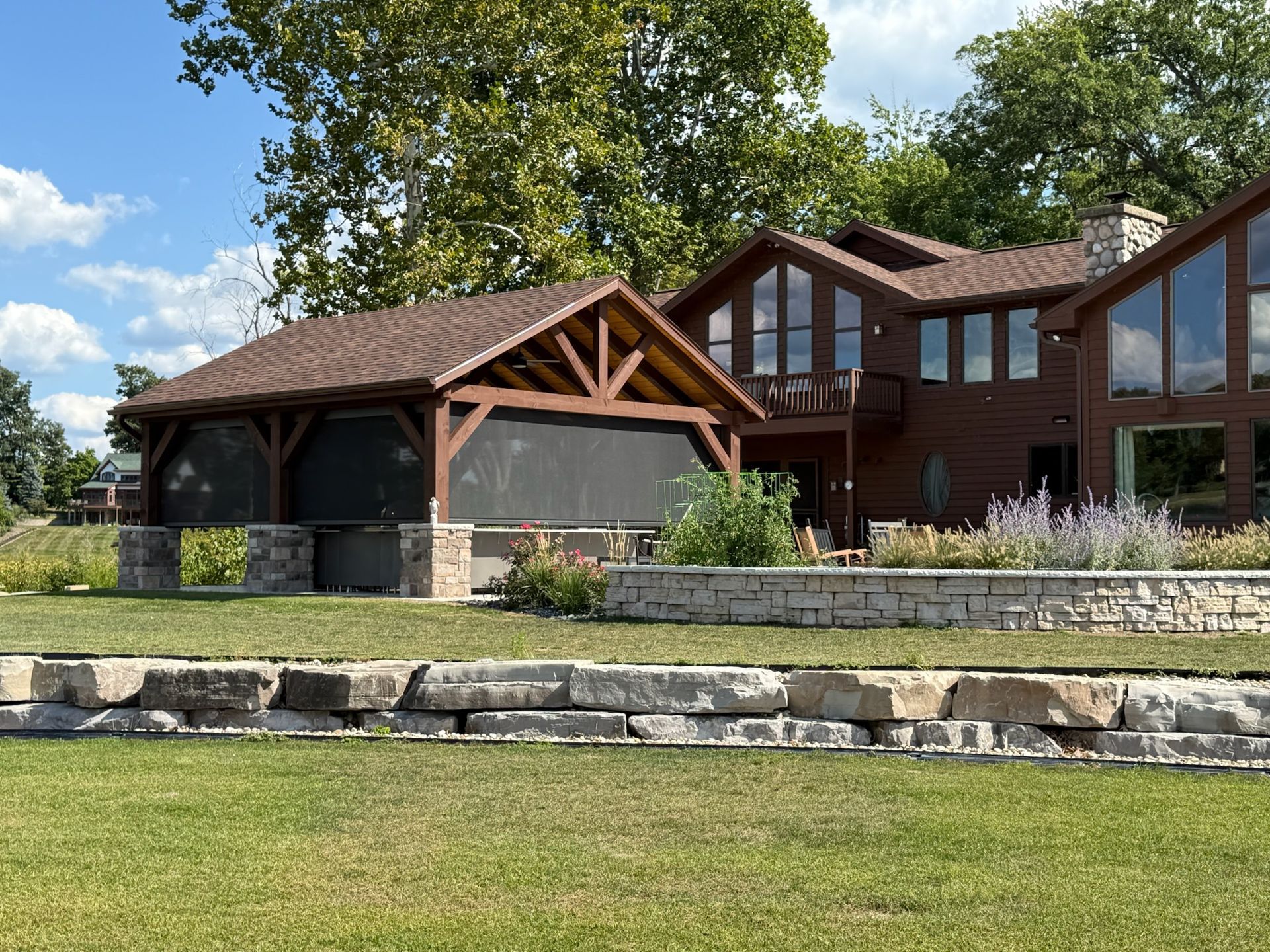 A brown wooden house with a screened-in timber pavilion stands on a grassy lawn with a tiered stone retaining wall.