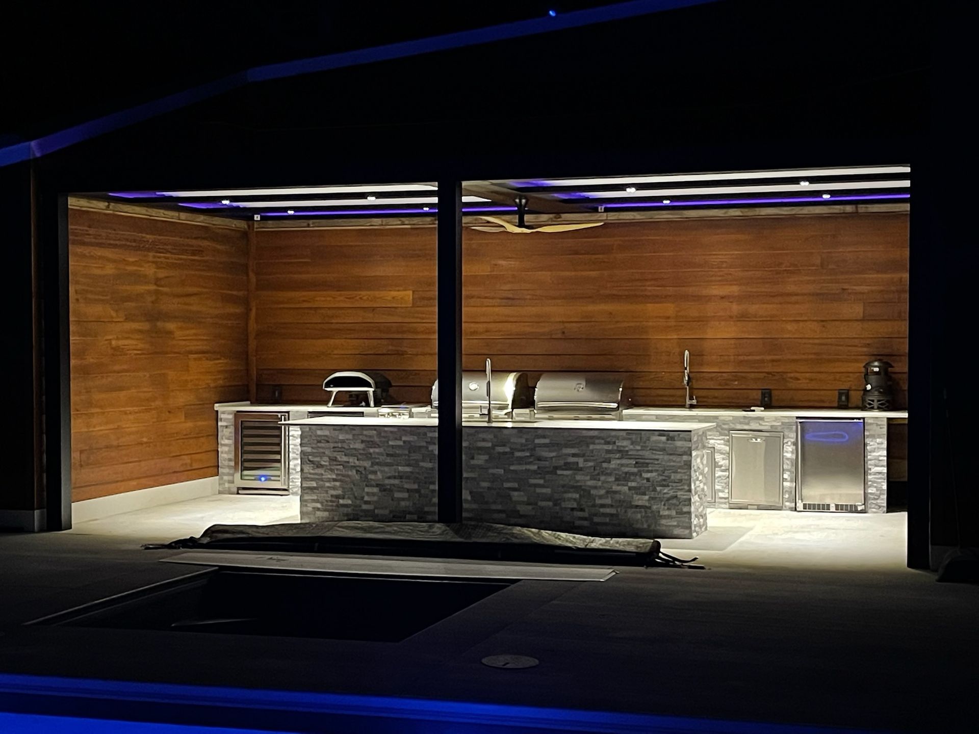 Modern outdoor kitchen with wood paneled walls, stone island, and stainless steel appliances illuminated at night.
