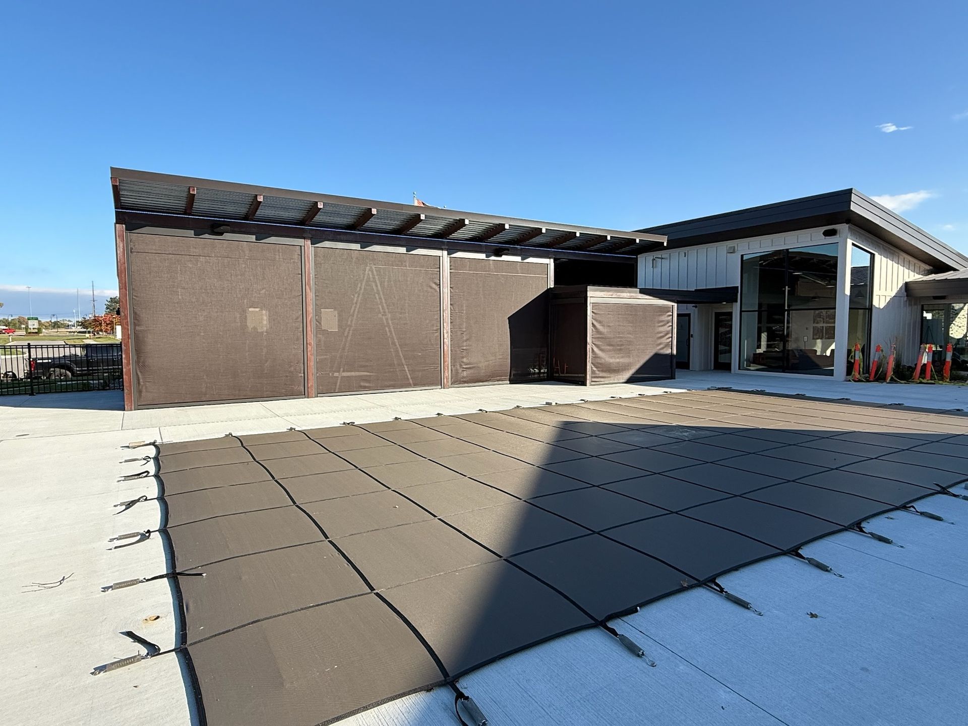 A grey safety cover over an outdoor pool next to a modern building with a covered patio area under a bright blue sky.