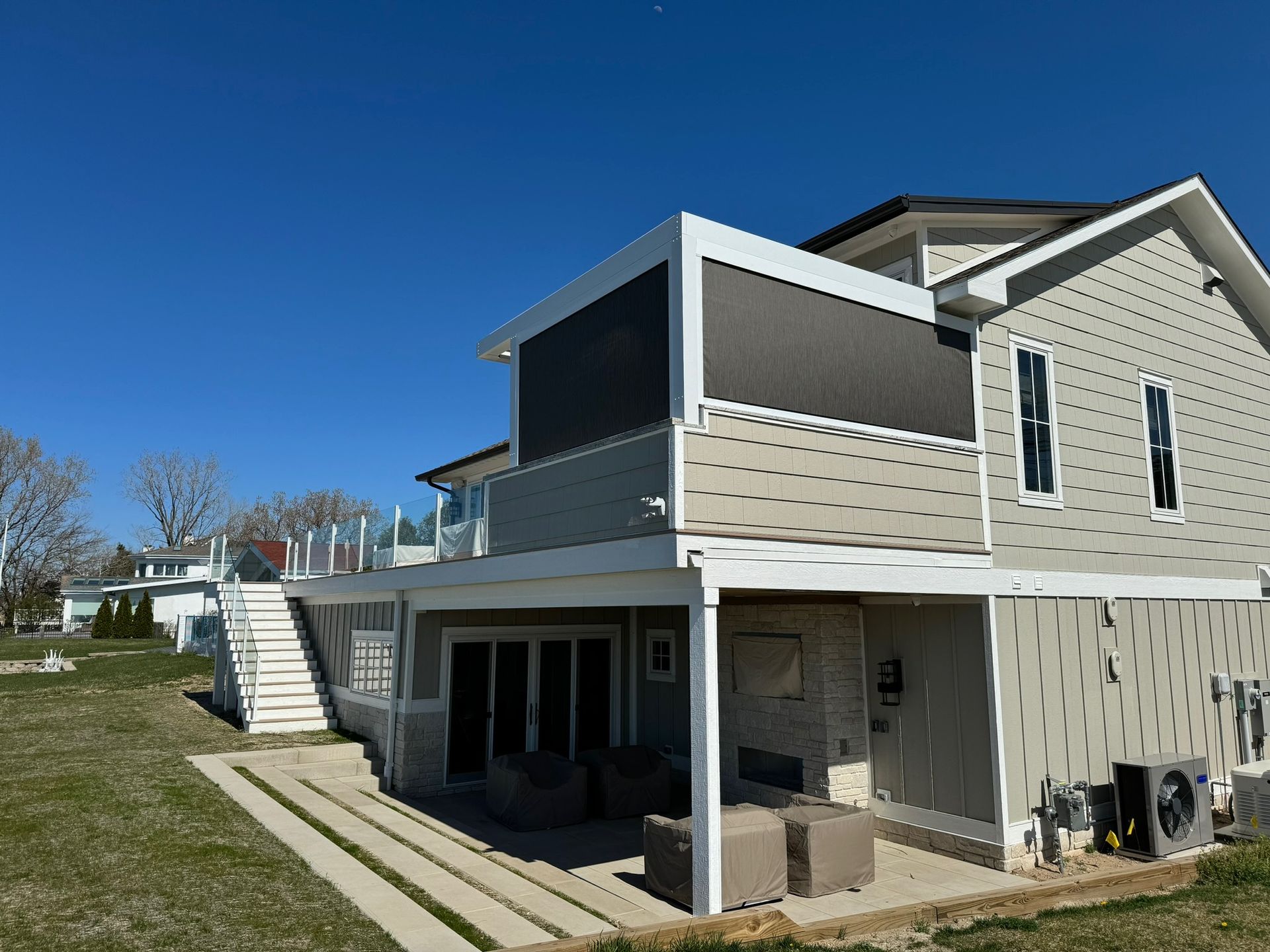 A two-story beige house with a covered lower patio, outdoor stairs, and a screened upper porch under a clear blue sky.