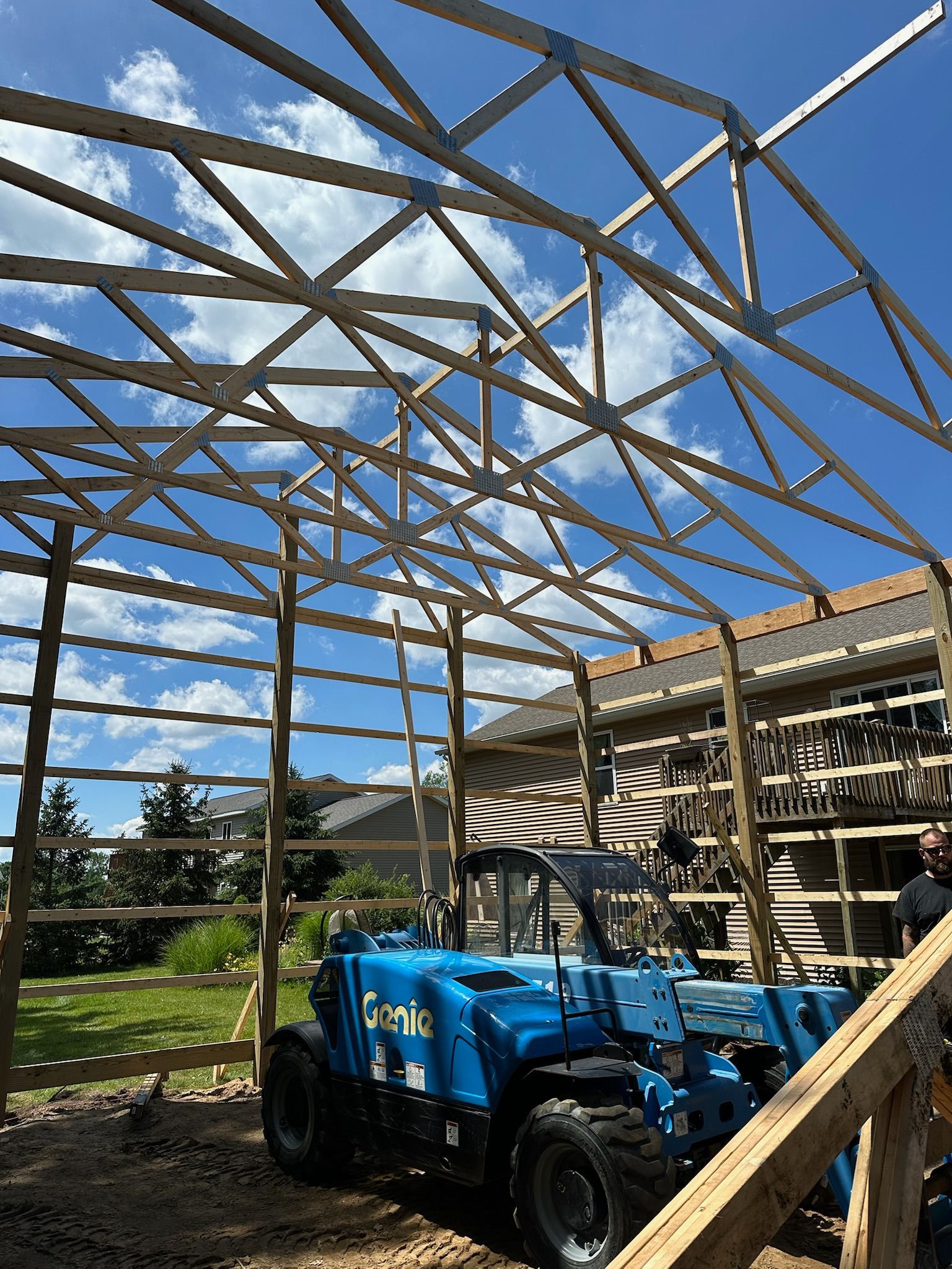 A blue telehandler sits under a wooden building frame construction in a residential yard under a sunny, blue sky.