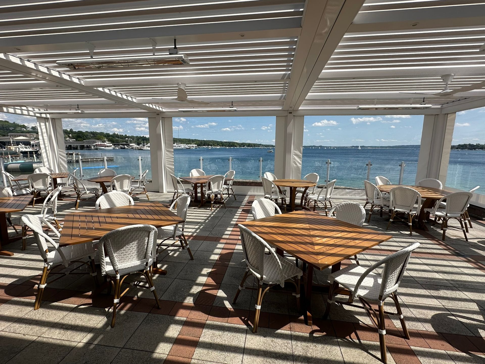 An outdoor restaurant patio with wooden tables and white chairs under a pergola, overlooking a sunny harbor.