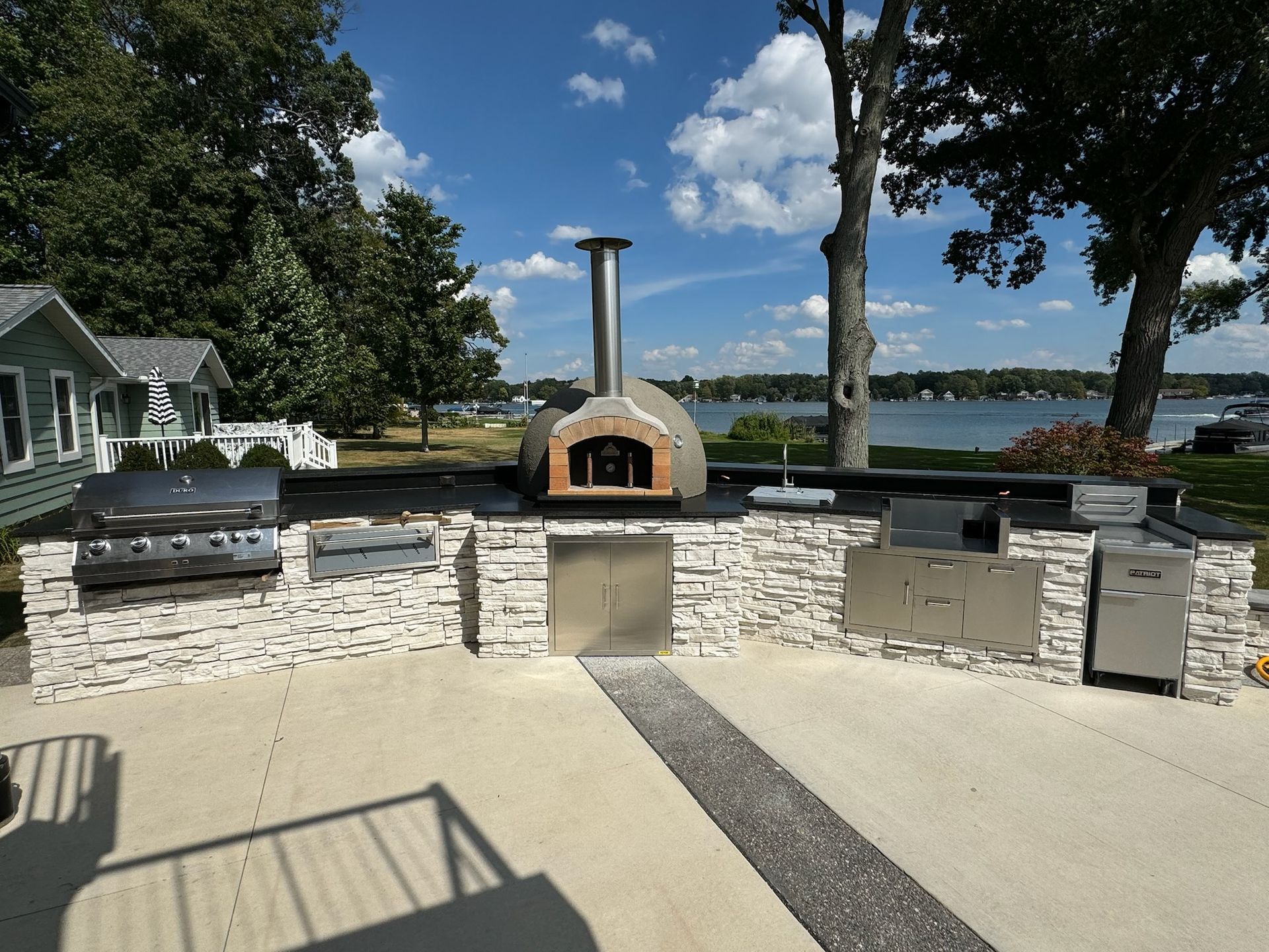 A large outdoor kitchen with stone masonry, a stainless steel pizza oven, and grills overlooking a lake under blue sky.