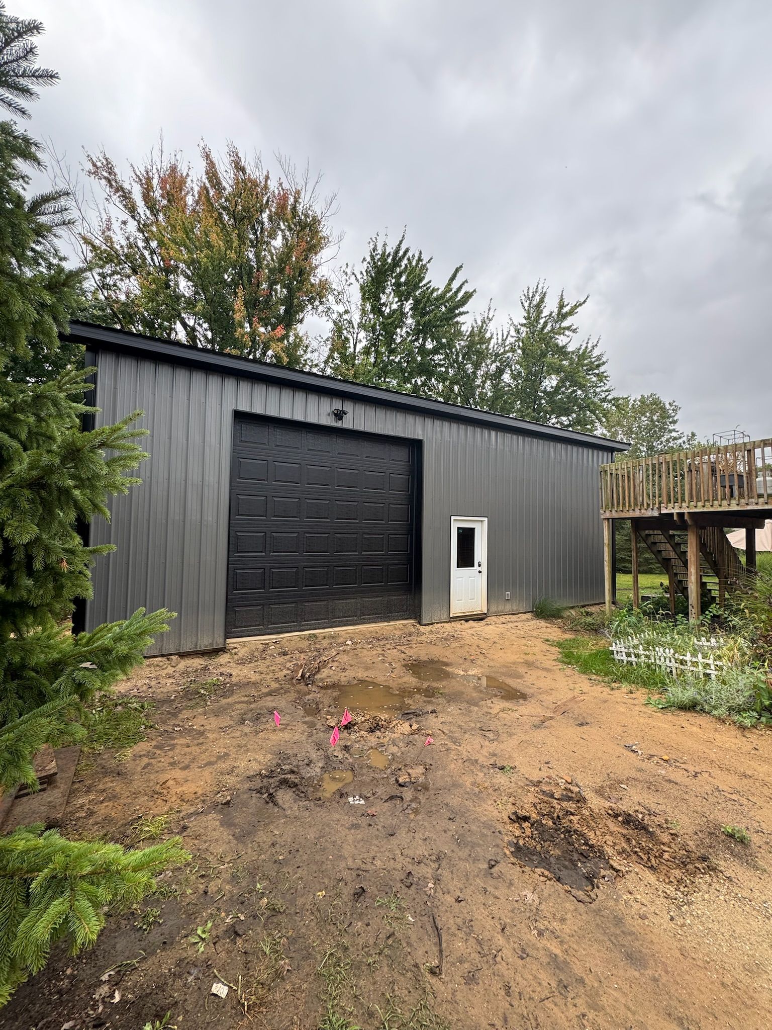 A gray metal shed with a black garage door and white entry door, set on a muddy lot near trees and a wooden deck.