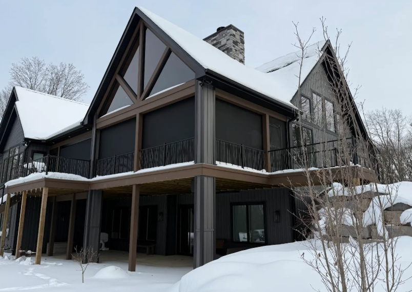 A dark-sided, multi-story home with a wrap-around deck and screened-in porch, surrounded by a deep winter snowpack.